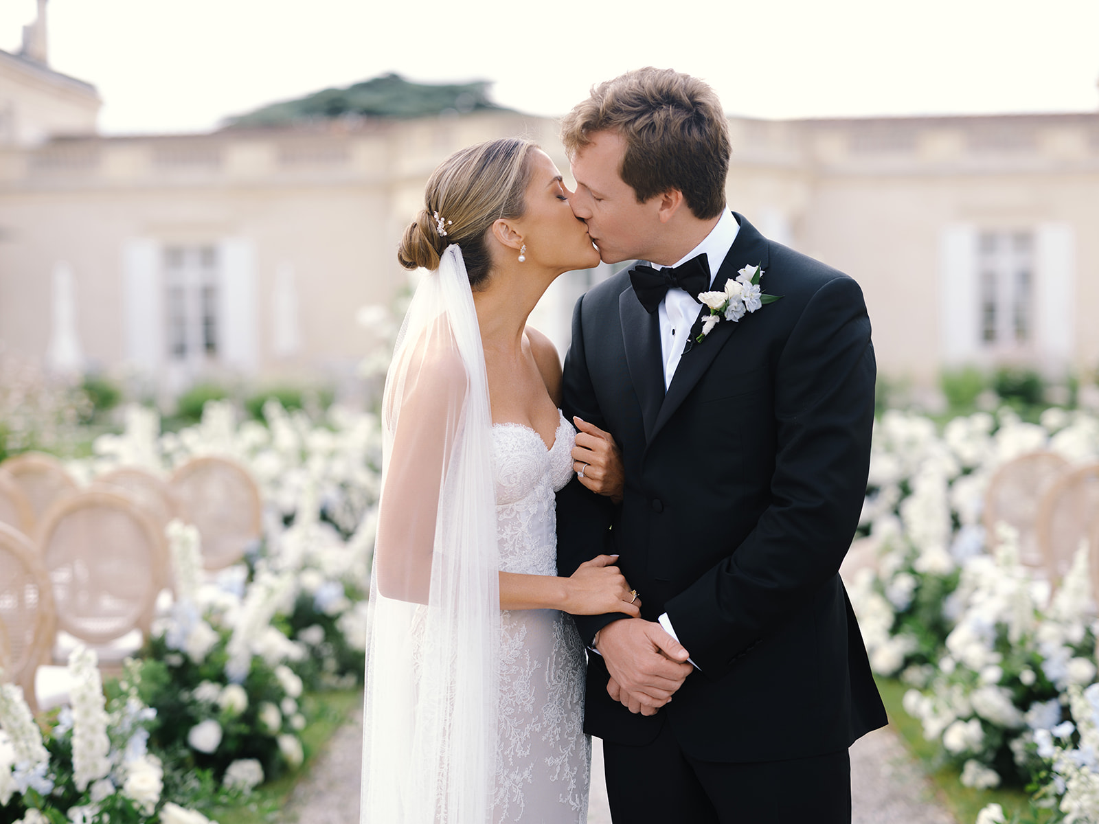 Bride and groom kissing in a classical chateau courtyard lined with white flowering plants