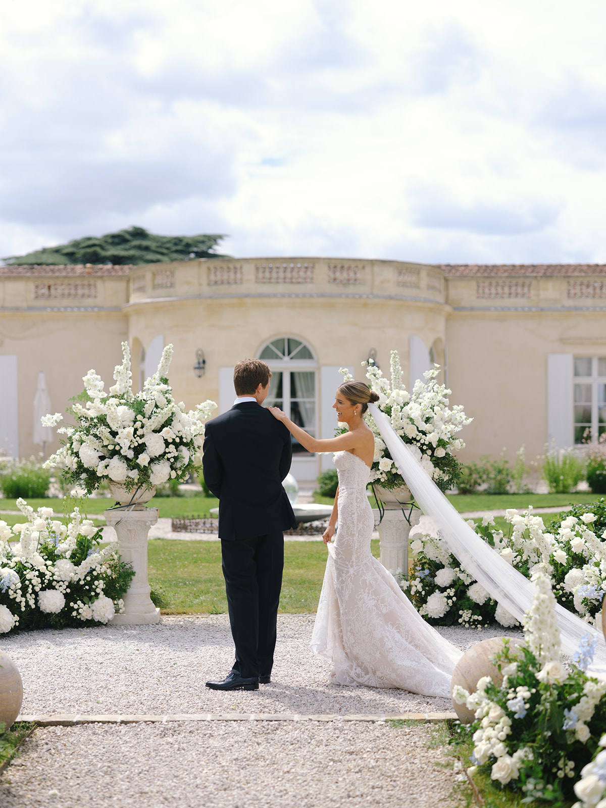 Couple at ceremony altar with white floral urns flanking gravel aisle before classical stone mansion