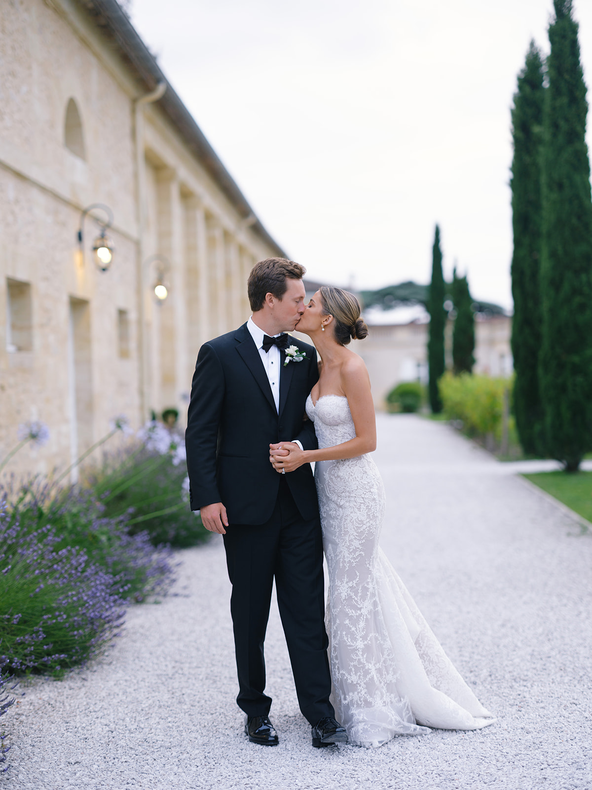 Bride and groom kissing on gravel path beside stone mansion with lavender and cypress trees