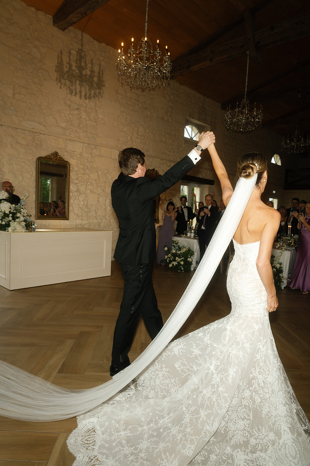 Bride and groom first dance in ballroom with crystal chandeliers, bride's lace train sweeping across parquet floor
