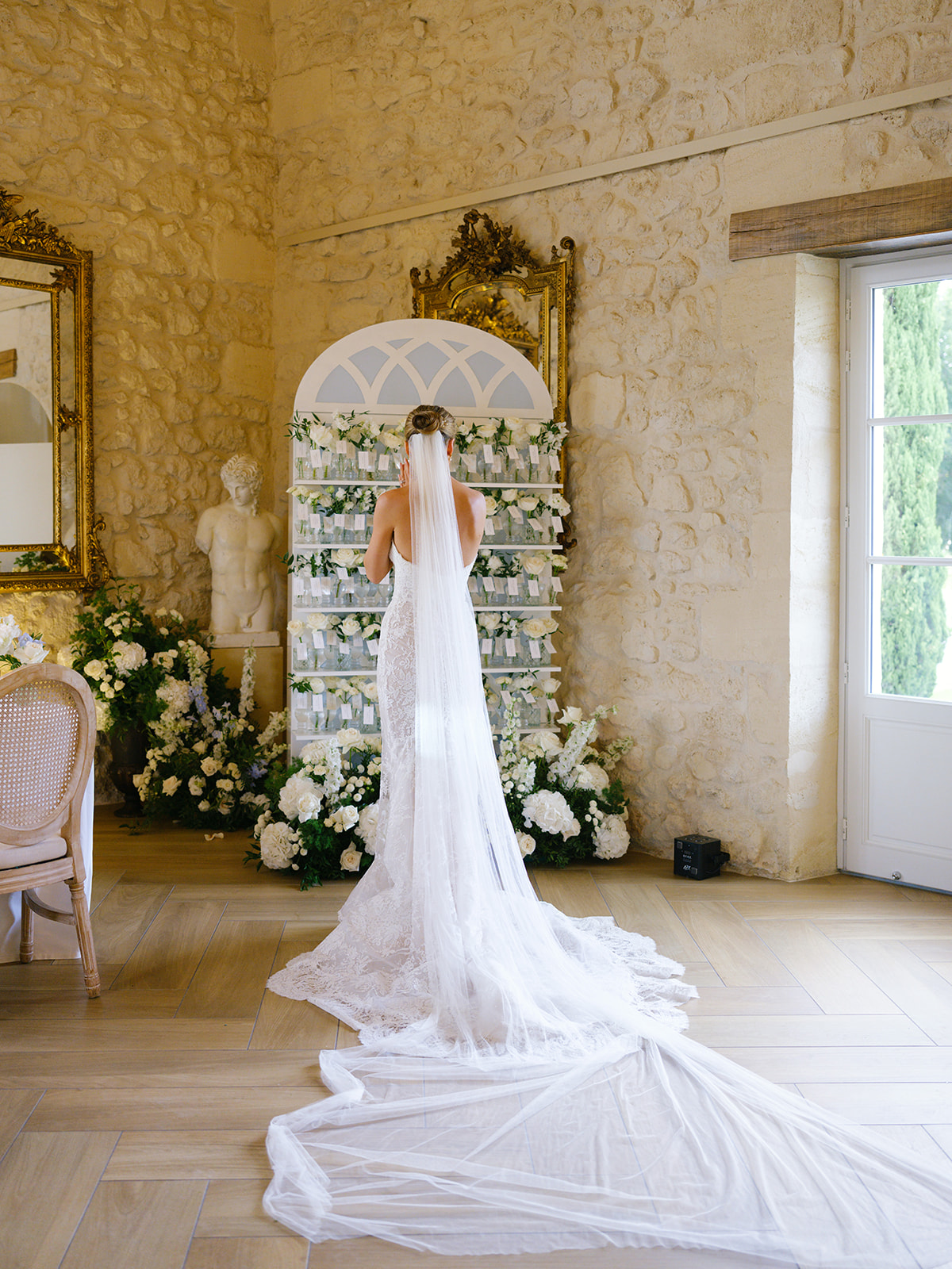 Bride in lace halter gown facing tiered flower vial installation with white roses and hydrangeas in classical interior