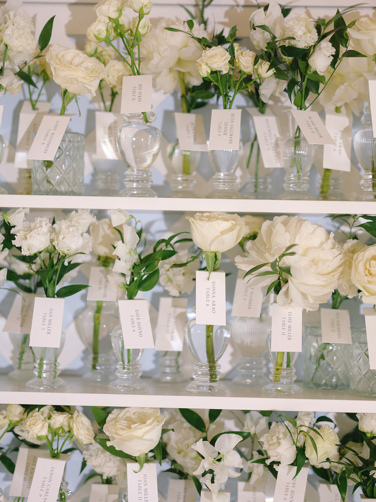 Escort card display with glass vases of cream roses and ivory carnations on white shelves, place cards beside each