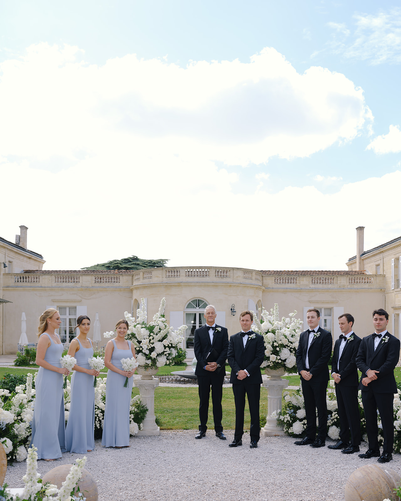 Bridal party posed in two lines at stone mansion courtyard with bridesmaids in blue dresses and groomsmen in black tuxedos