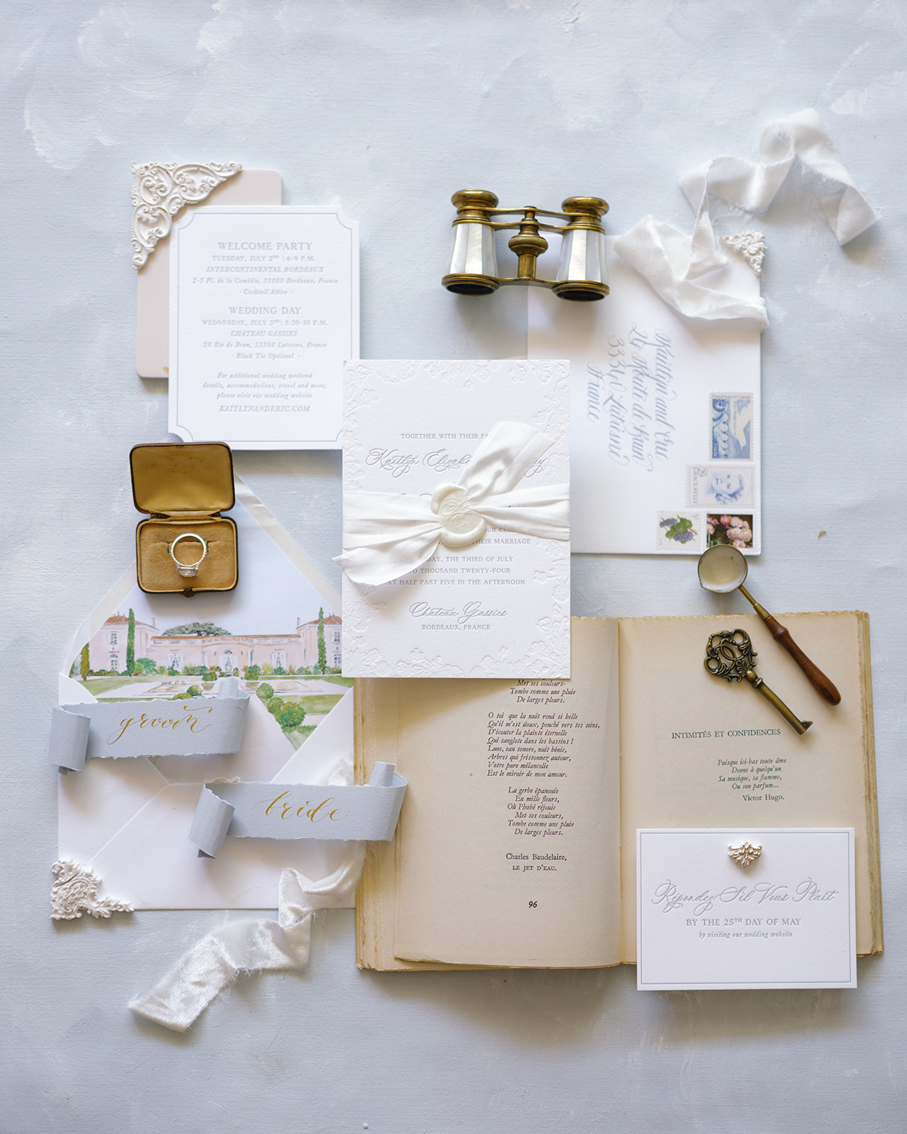 Wedding stationery flat lay with calligraphy invitation, château illustration, ring in brass box, and vintage opera binoculars