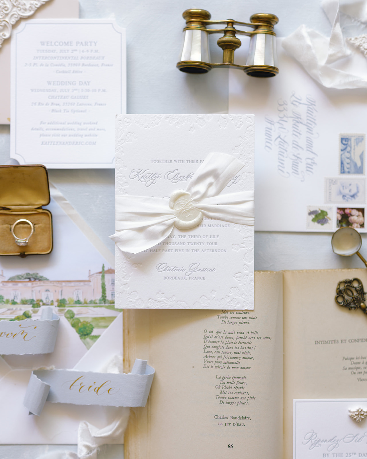 Wedding stationery flat lay with embossed invitation, gold ring box, vintage binoculars, and calligraphy