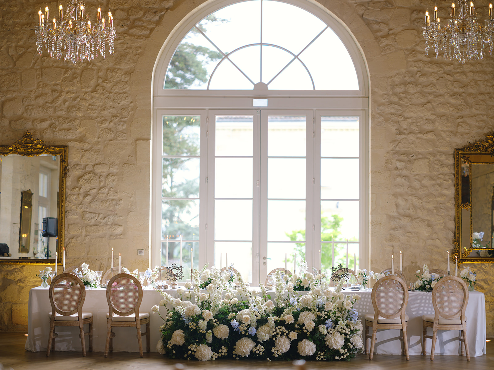 Long reception table with white hydrangea floral runner and candles in a classical stone ballroom with arched window