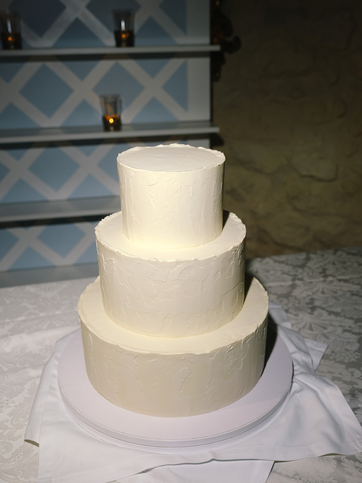 Three-tiered ivory buttercream wedding cake on glass stand with geometric backdrop