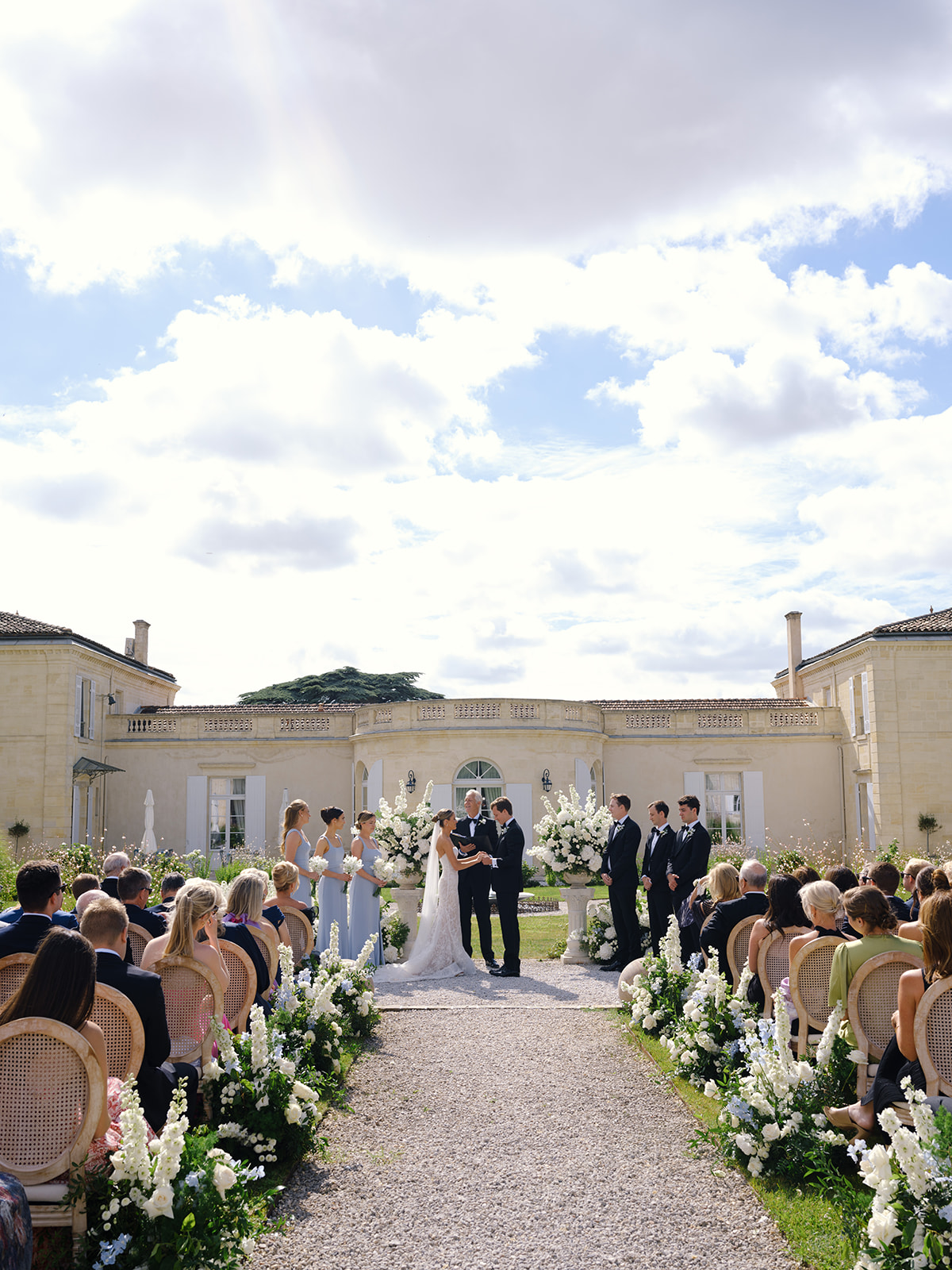 Ceremony in classical stone courtyard with bridesmaids in light blue, groomsmen in dark suits and white floral aisle