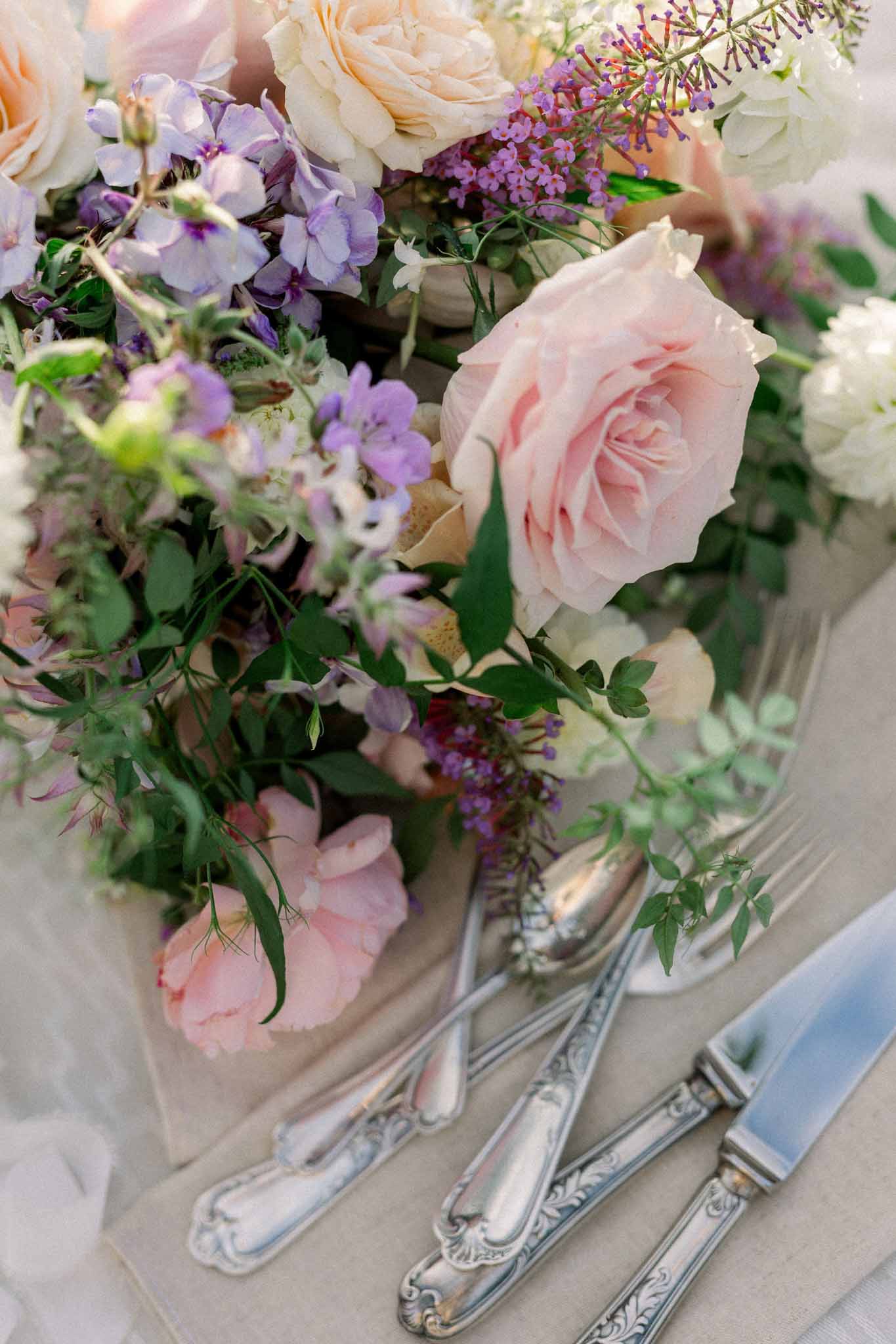 Close-up of blush roses, peach garden roses, and purple sweet peas beside ornate silver flatware