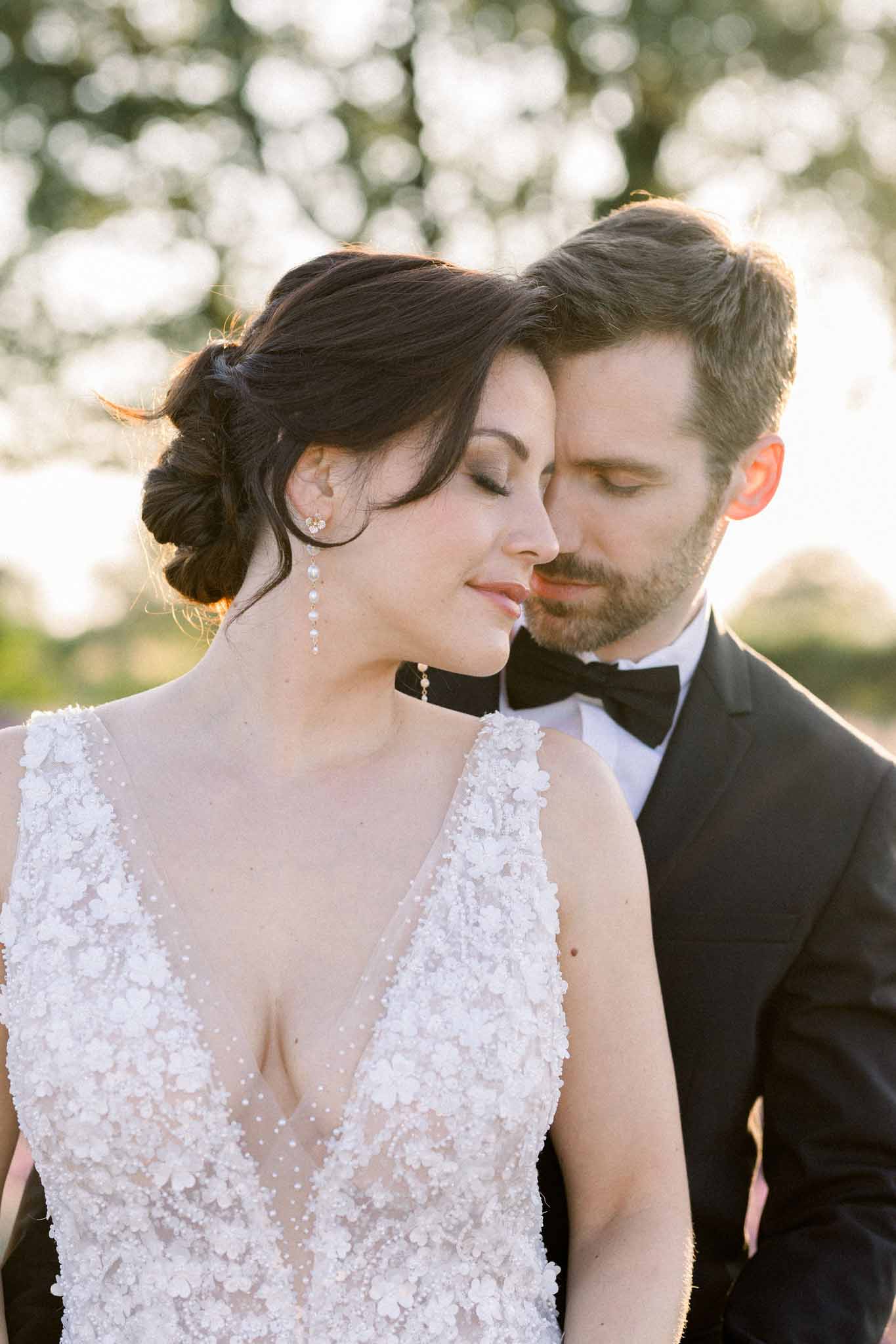 Close-up couple portrait at golden hour, bride in ivory lace gown and groom in tuxedo with eyes closed