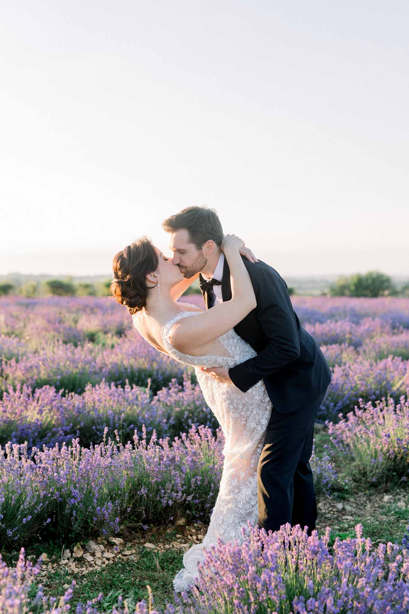 Bride and groom sharing a dip kiss in a blooming lavender field at golden hour with distant hills
