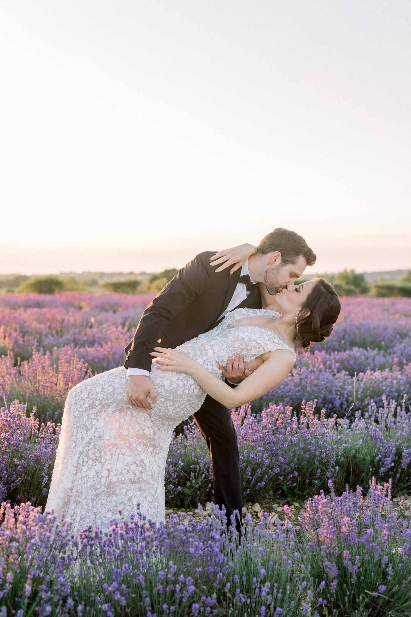 Groom dipping bride in ivory lace gown for a kiss in a lavender field at golden hour