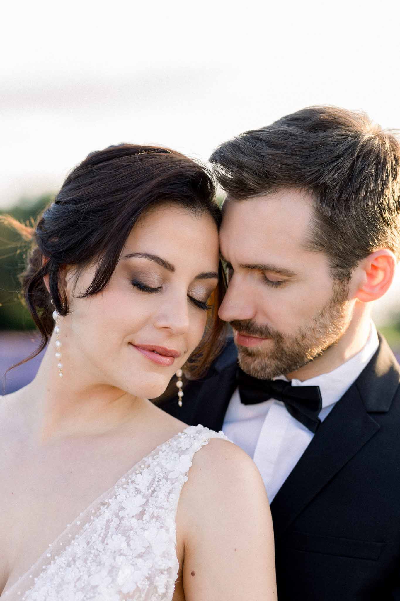 Bride in beaded dress and groom in black tuxedo in tender close-up portrait outdoors