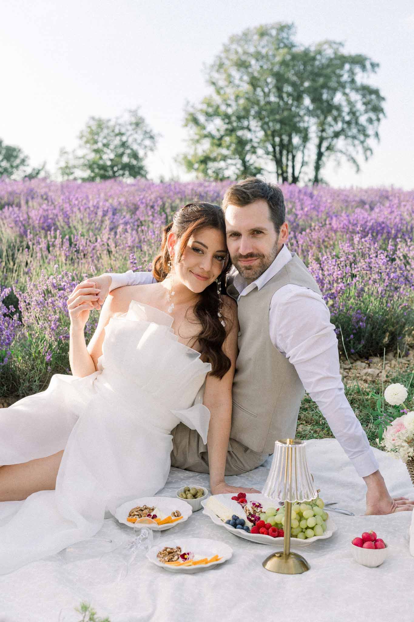 Bride and groom seated at white-draped table with fruit bowls in lavender field during portrait session
