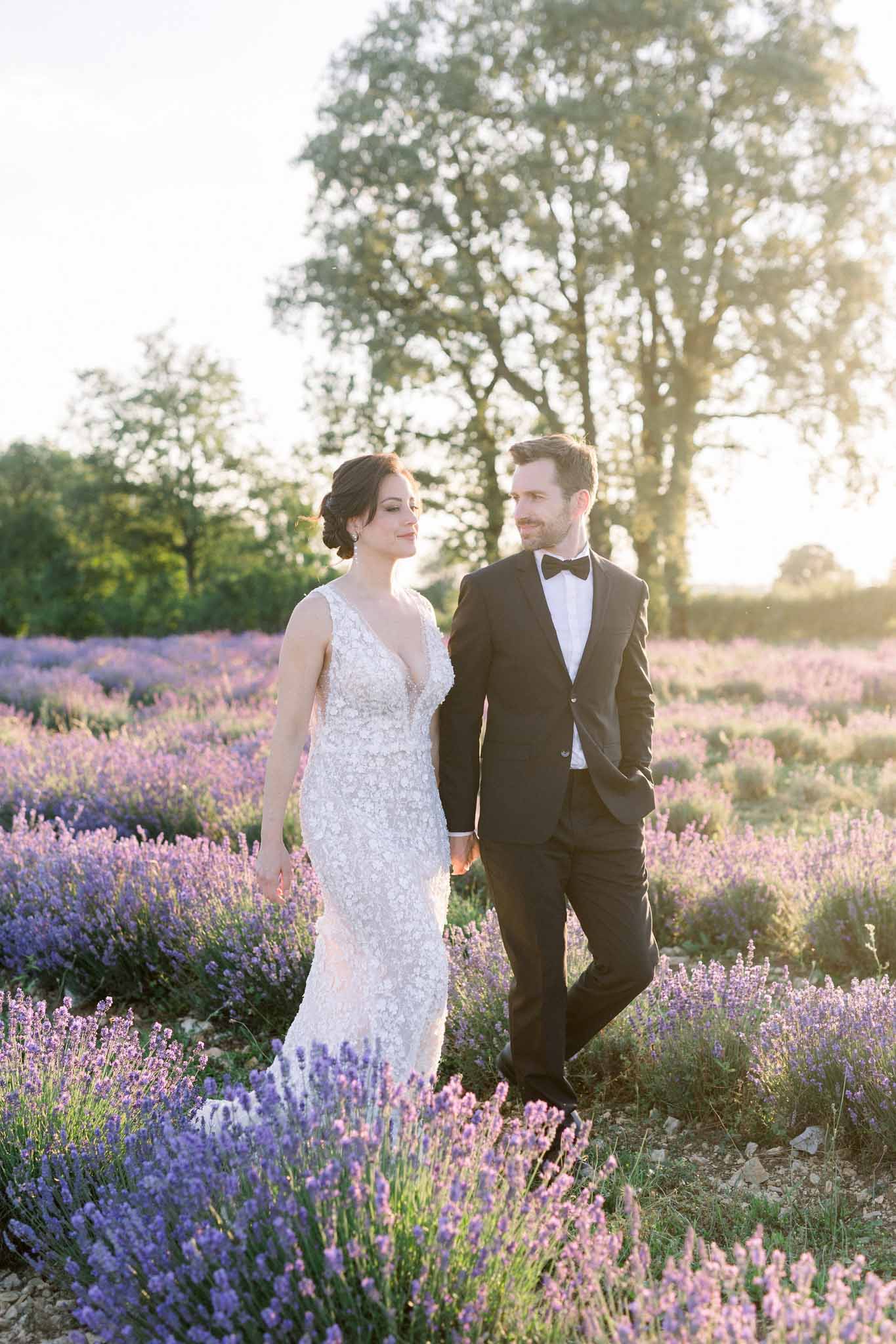 Bride and groom walking hand-in-hand through blooming lavender field at golden hour, framed by tall trees on the horizon