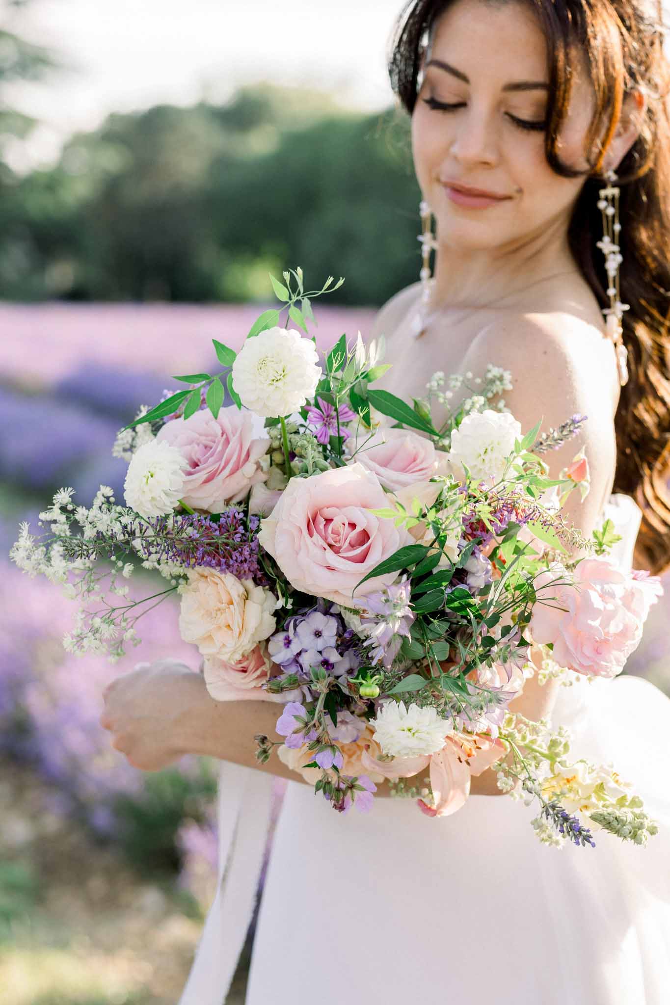 Bride holding lush bouquet of blush roses, ivory peonies, and purple sweet pea in a lavender field