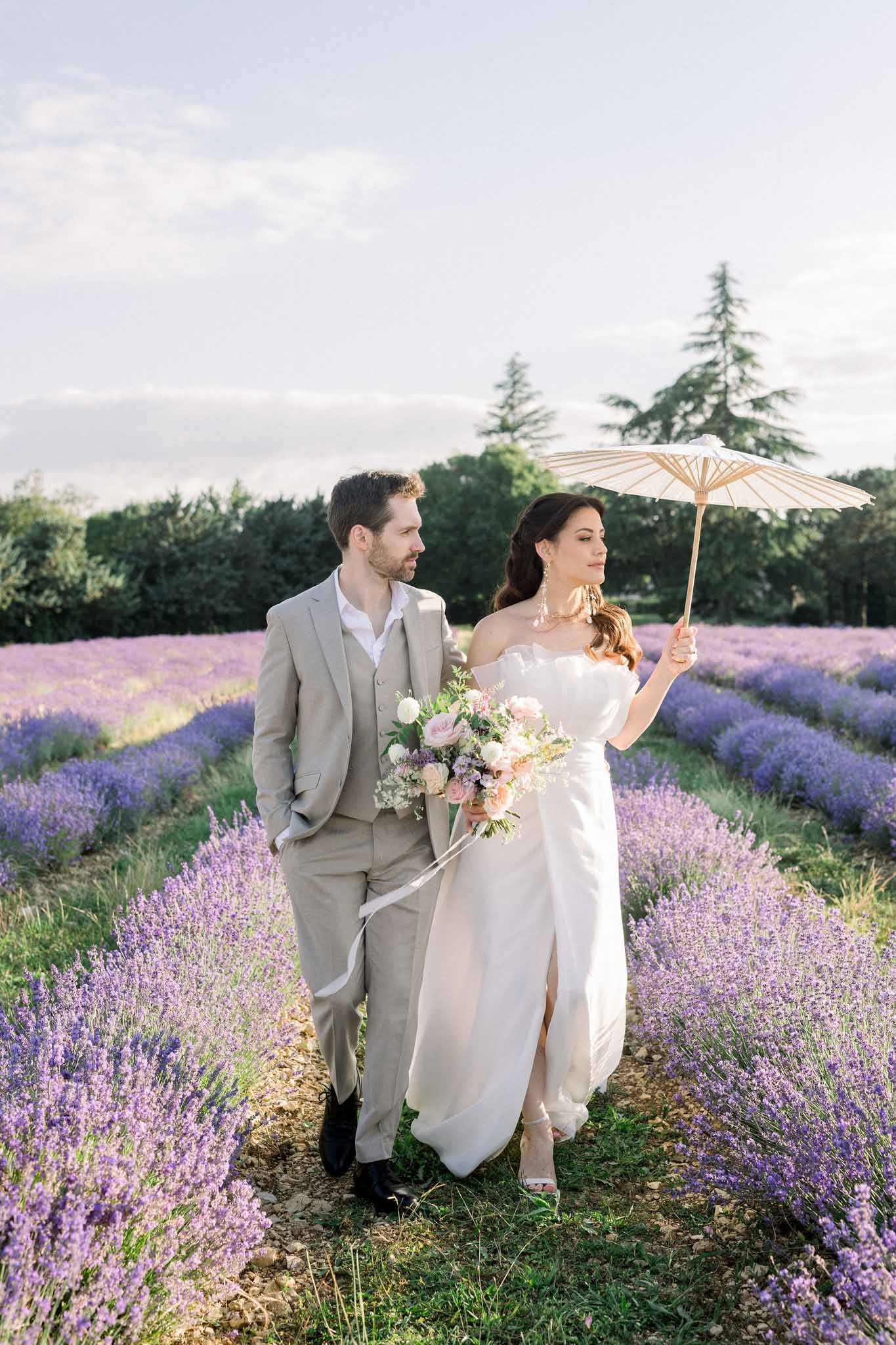 Bride with parasol and groom in tan suit walking through blooming lavender field in late afternoon light