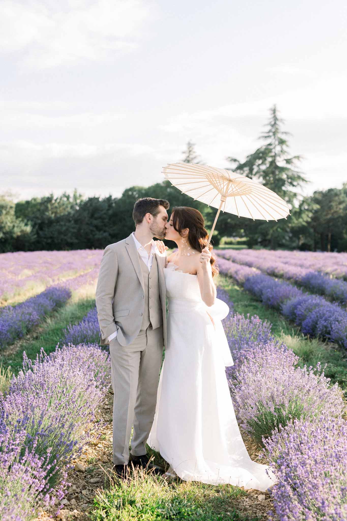 Bride and groom kissing under cream striped parasol in lavender field, groom in light taupe three-piece suit