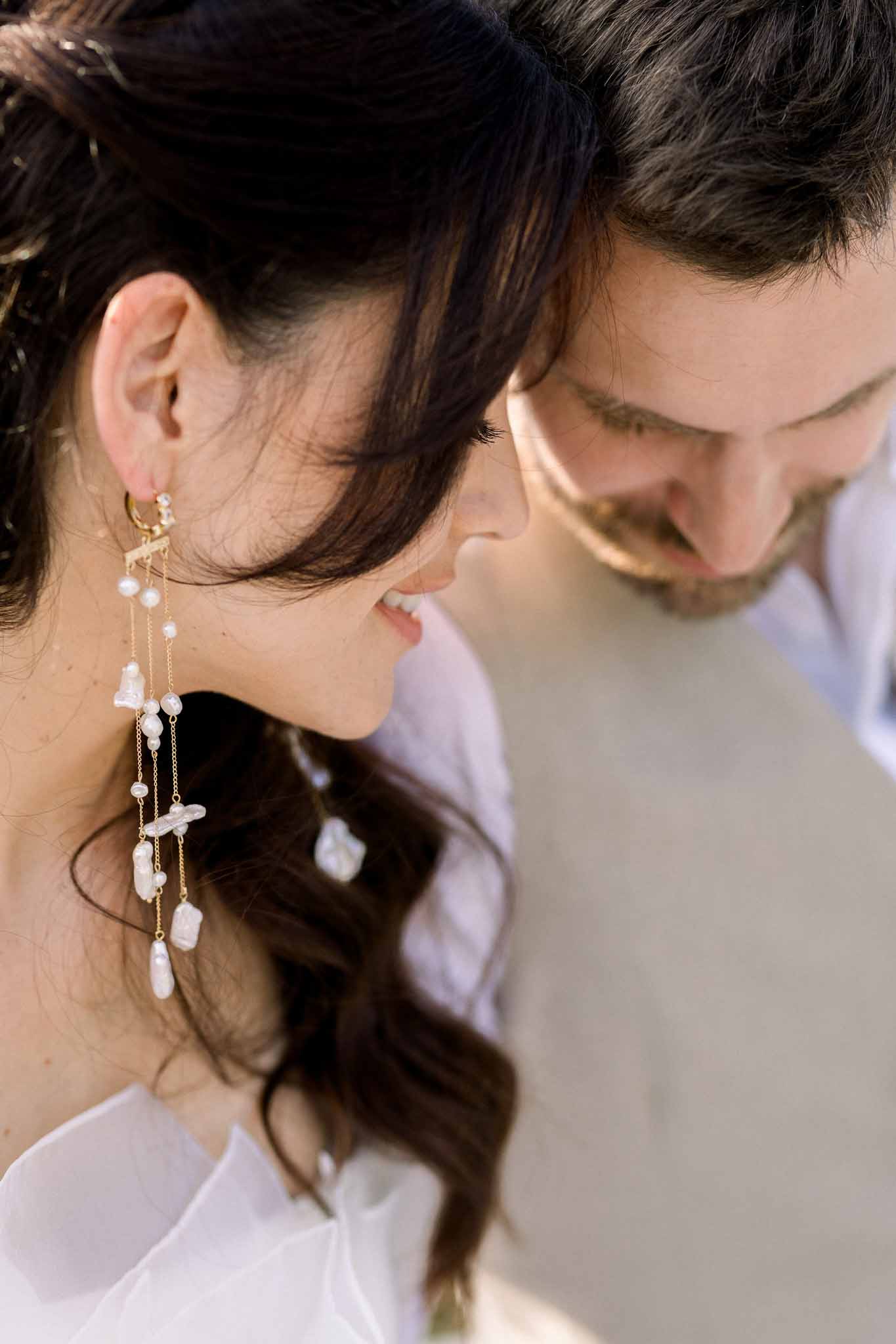 Overhead close-up of bride's baroque pearl drop earrings with groom softly blurred behind her