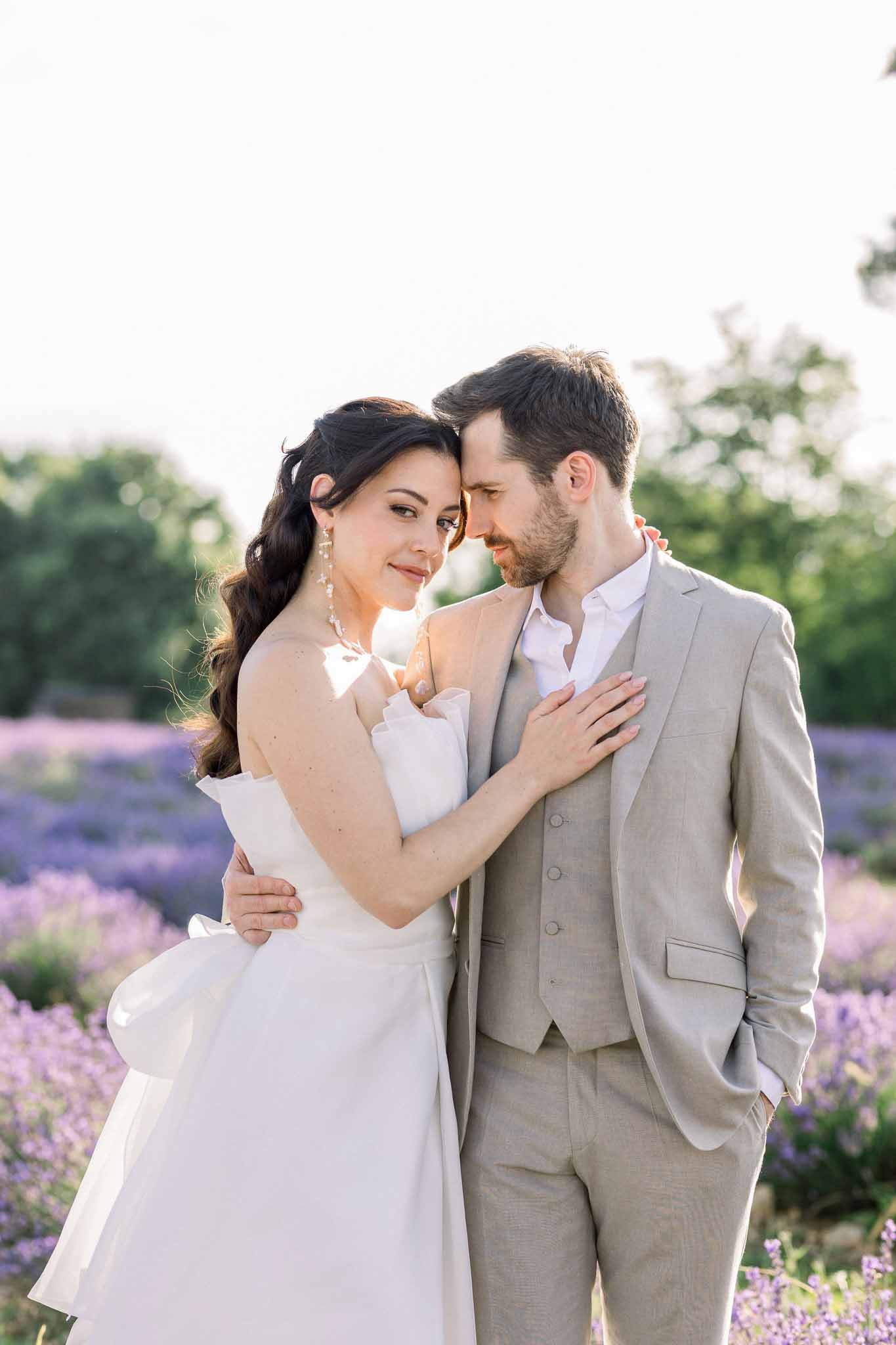 Bride and groom in lavender field, bride in ivory A-line dress with pearl earrings, groom in beige linen suit