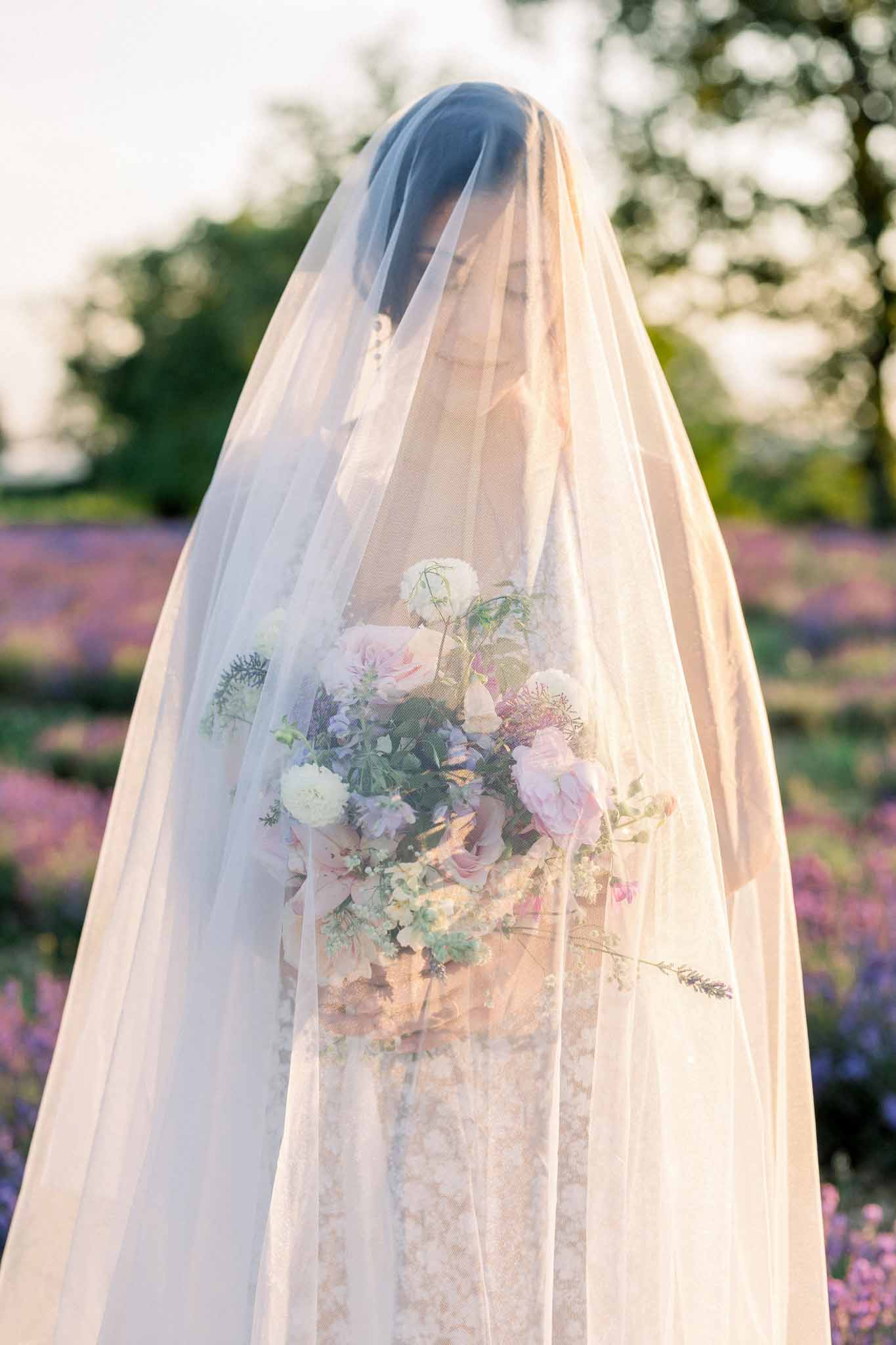 Bride from behind in ivory lace gown and flowing veil holding pink rose and purple scabiosa bouquet in garden