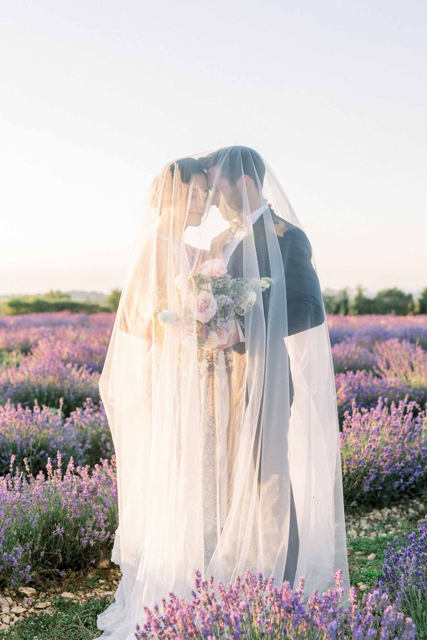 Bride and groom share an intimate moment under a sheer veil in a lavender field with golden backlight