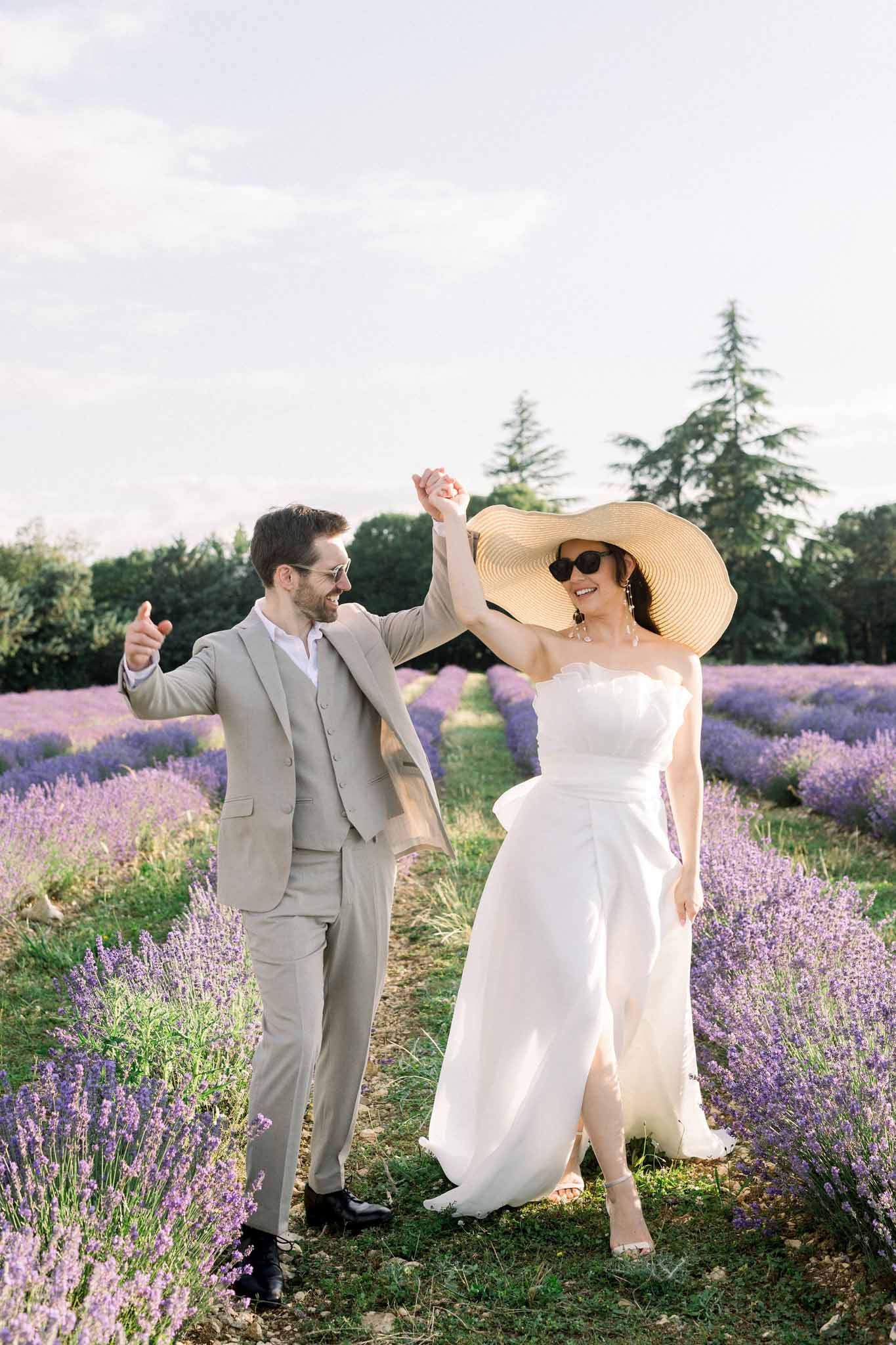 Bride in white off-shoulder gown with straw hat and groom in taupe linen suit celebrate in lavender field