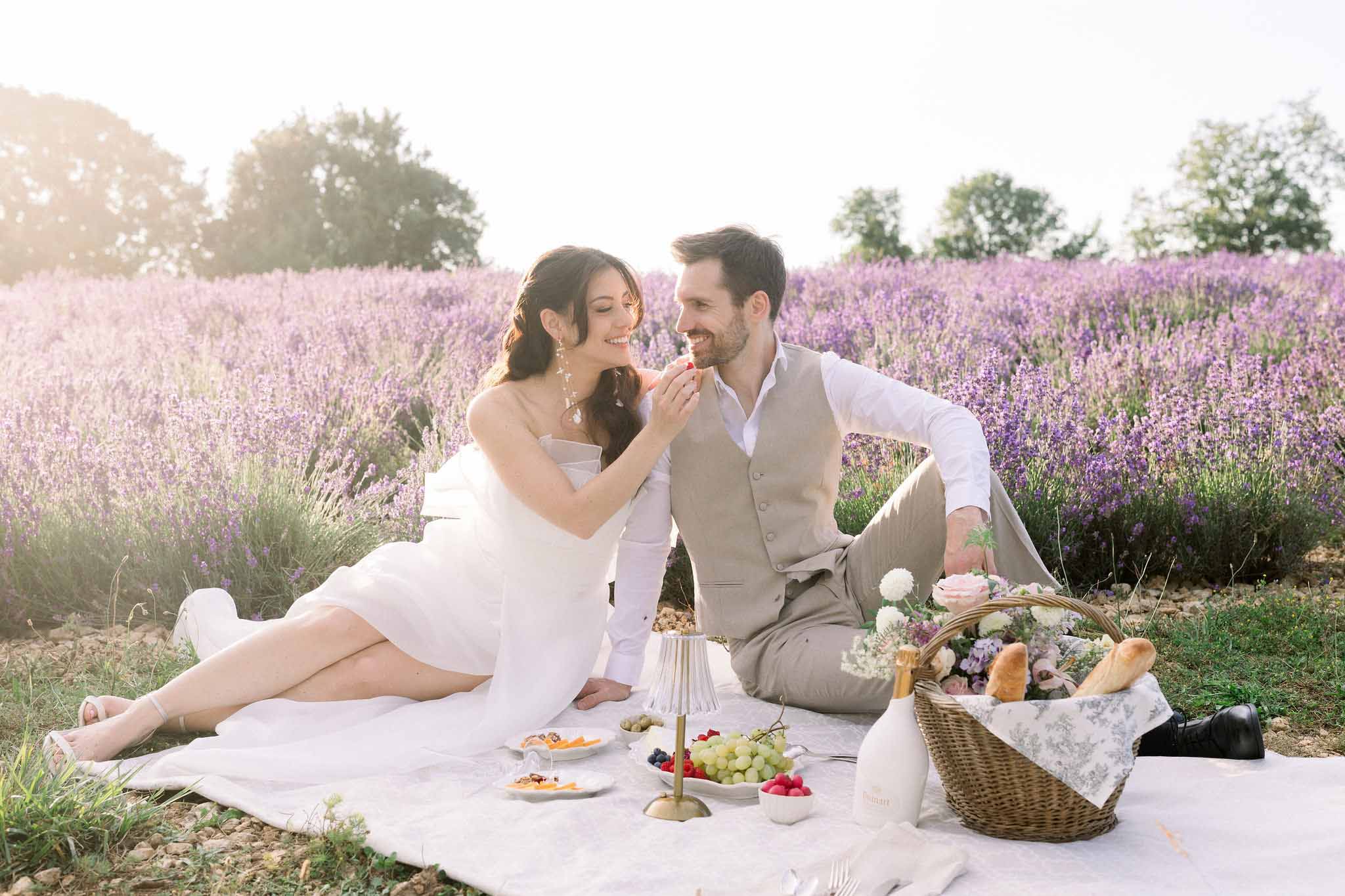 Bride and groom sharing a picnic on a white blanket in a lavender field during golden hour