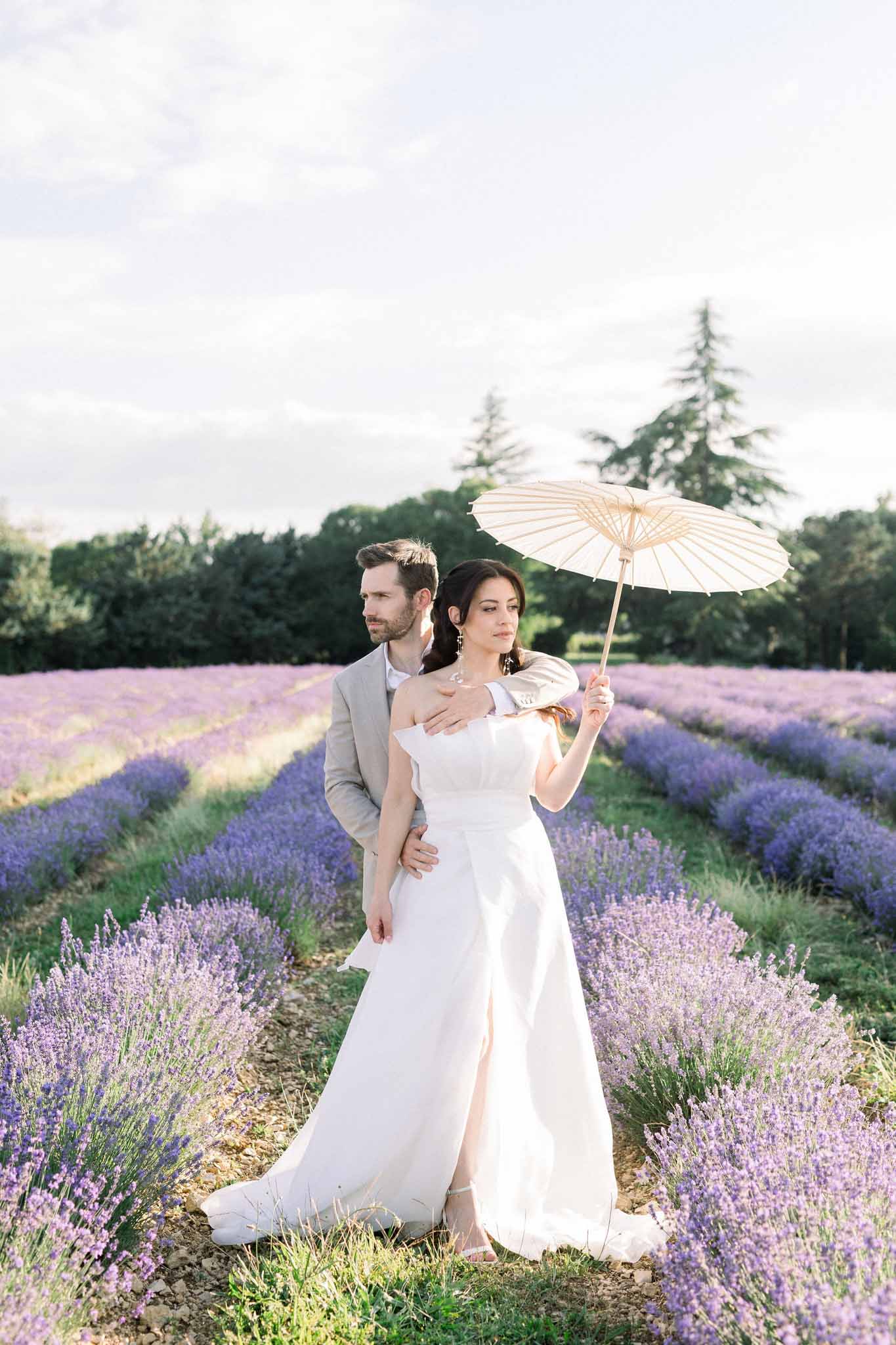Bride and groom posing in lavender field, bride holding cream parasol, groom in grey suit