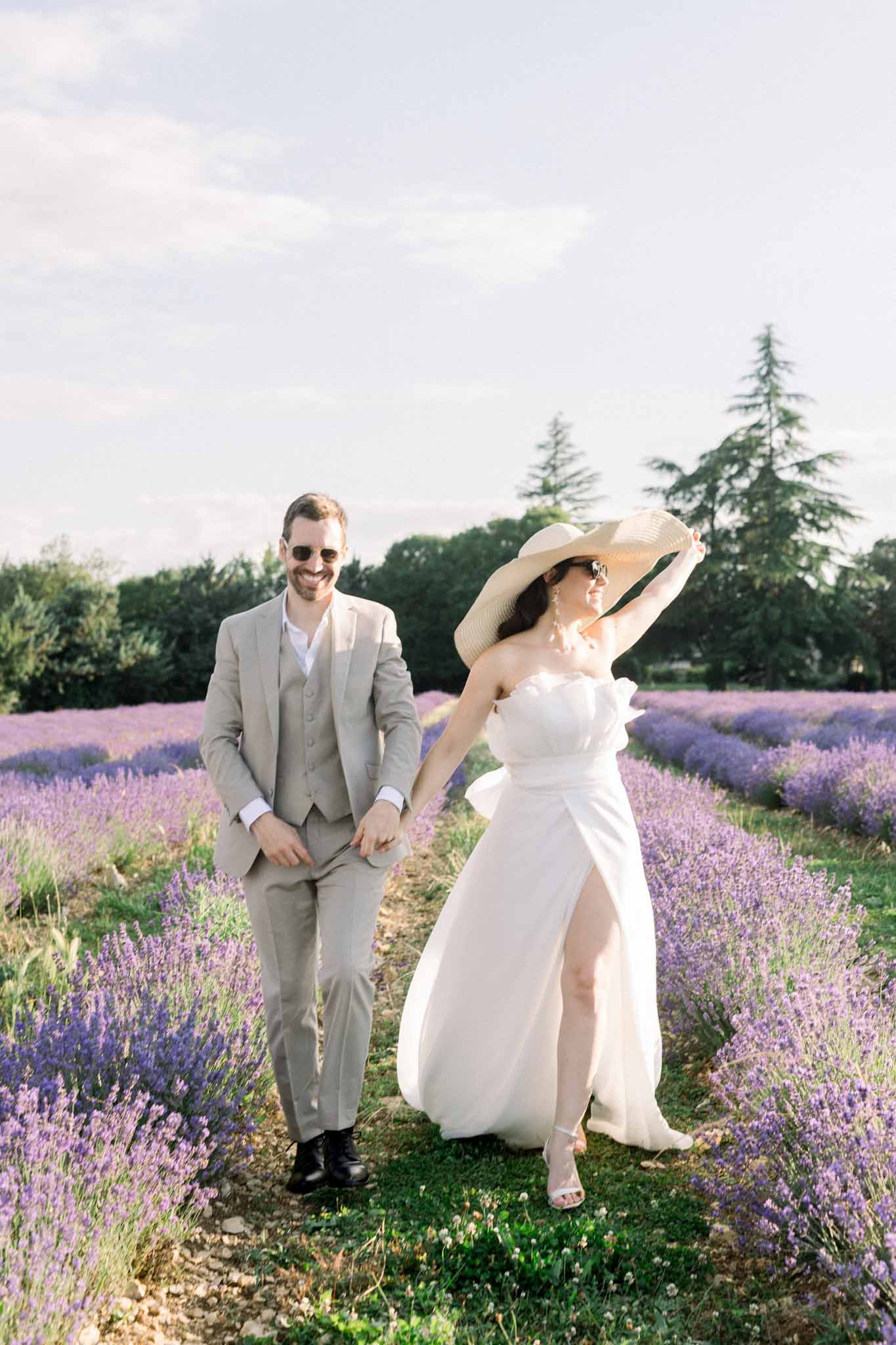 Bride in ivory strapless gown with wide-brim straw hat and groom in light gray suit walk through lavender field in full bloom
