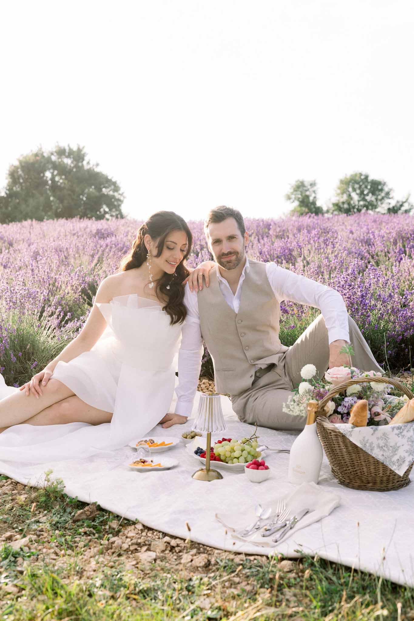 Bride and groom on cream linen picnic blanket in lavender field with wicker baskets and floral arrangements