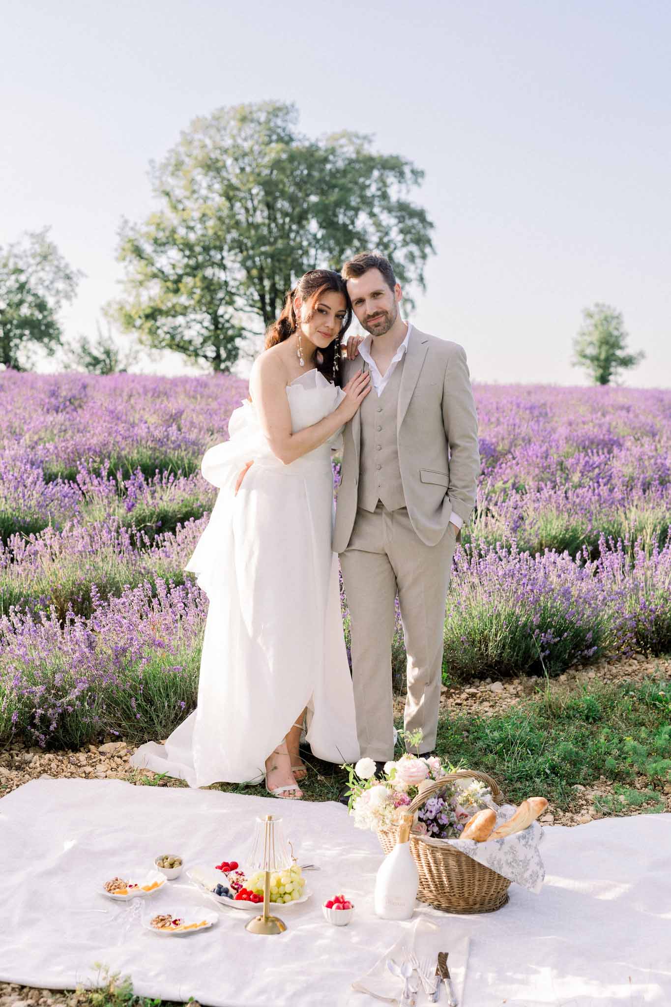 Bride in ivory gown and groom in taupe linen suit standing behind picnic spread in a lavender field