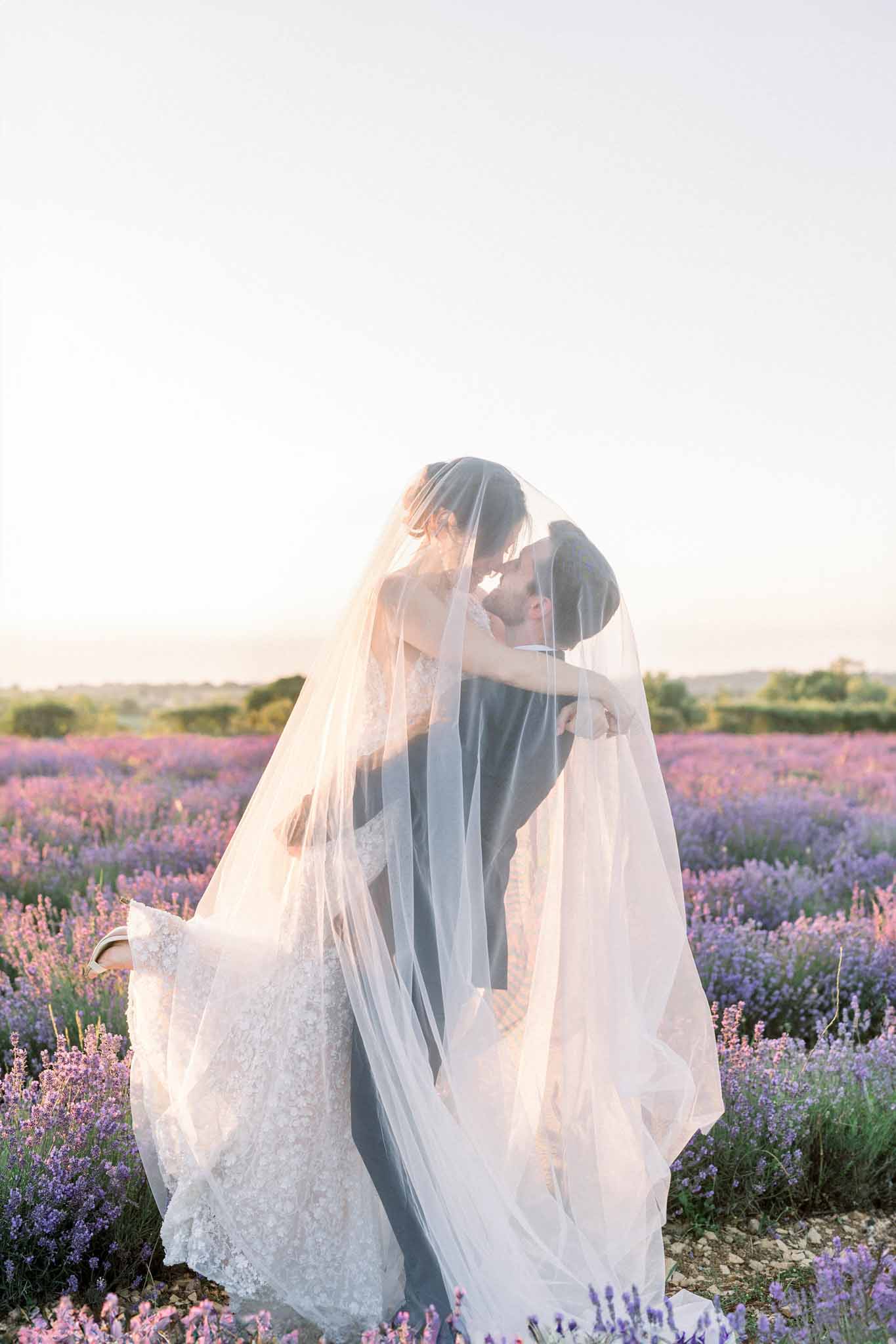 Bride and groom in lavender field, white lace dress with long ivory veil, golden hour wide portrait