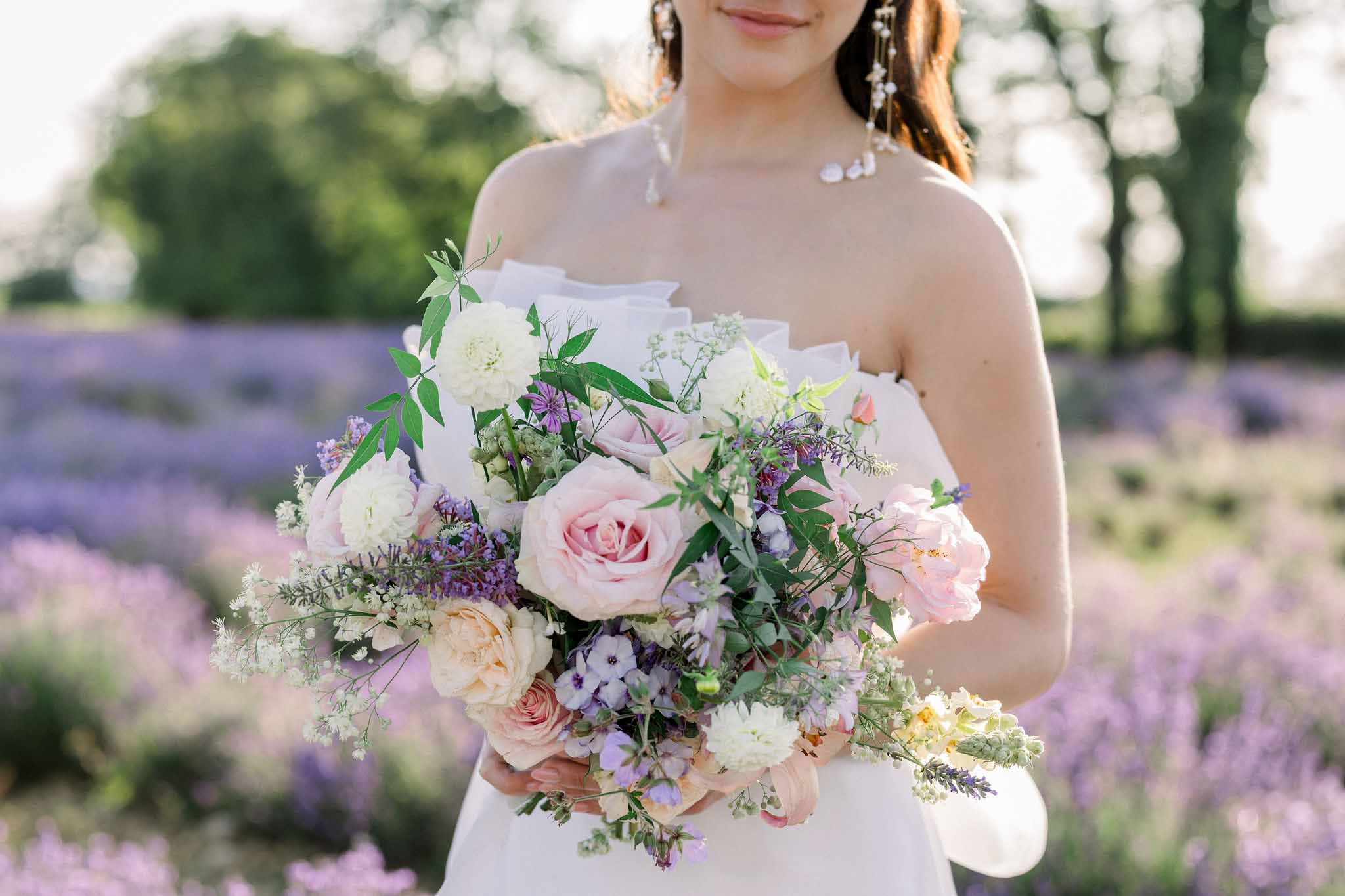 Bride holding bouquet of blush roses, ivory peonies and purple statice in lavender field, waist-up portrait