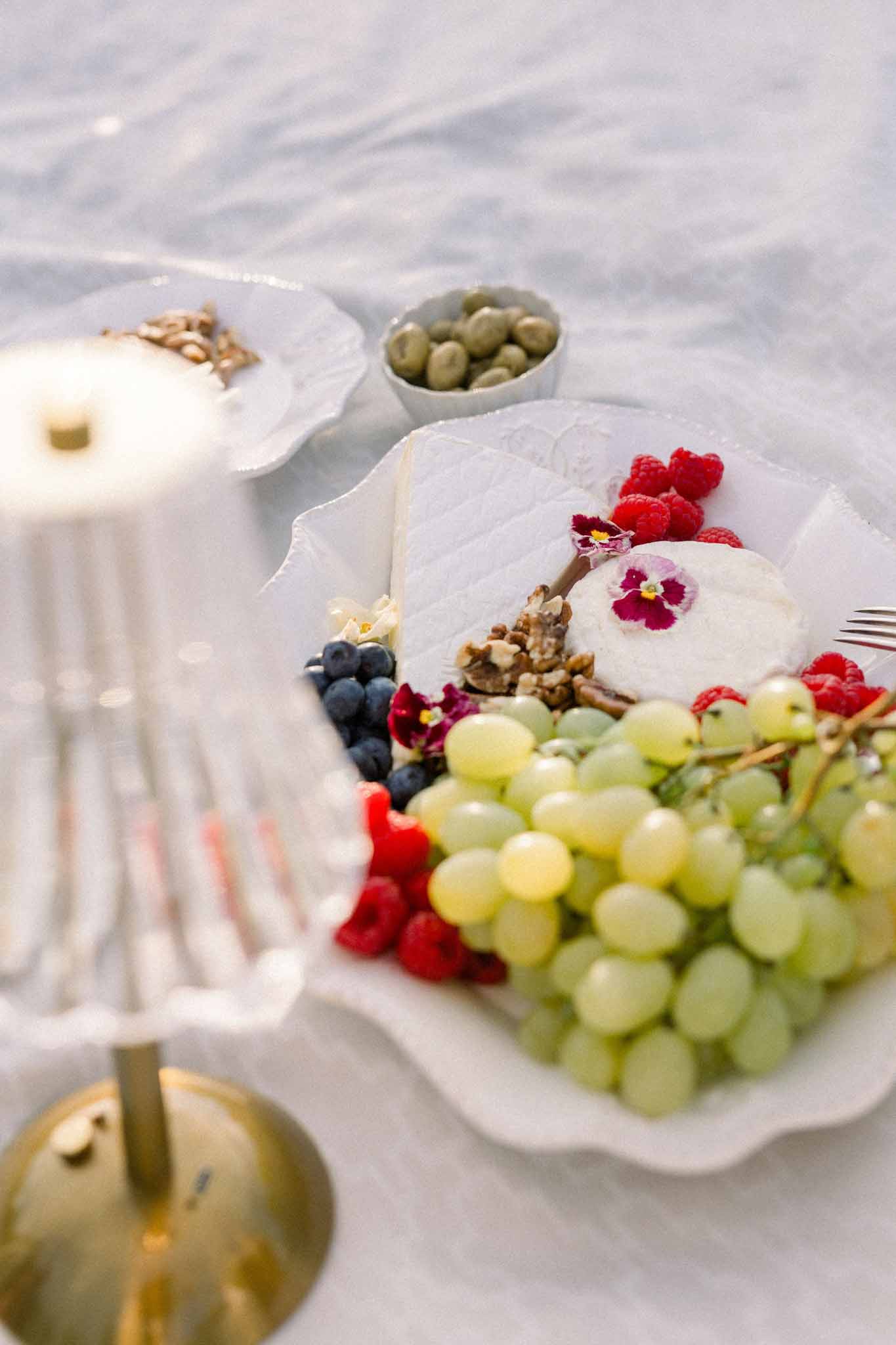 Close-up of shell-shaped serving dishes with grapes, blueberries, raspberries, walnuts, olives, and edible flowers on white linen