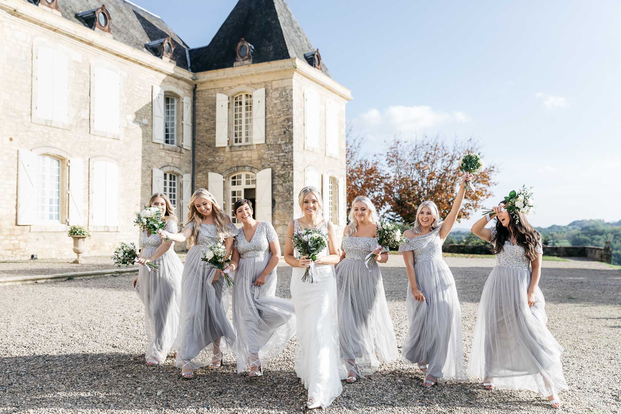 Bride in ivory lace gown walking with six bridesmaids in silver-grey tulle dresses on chateau forecourt