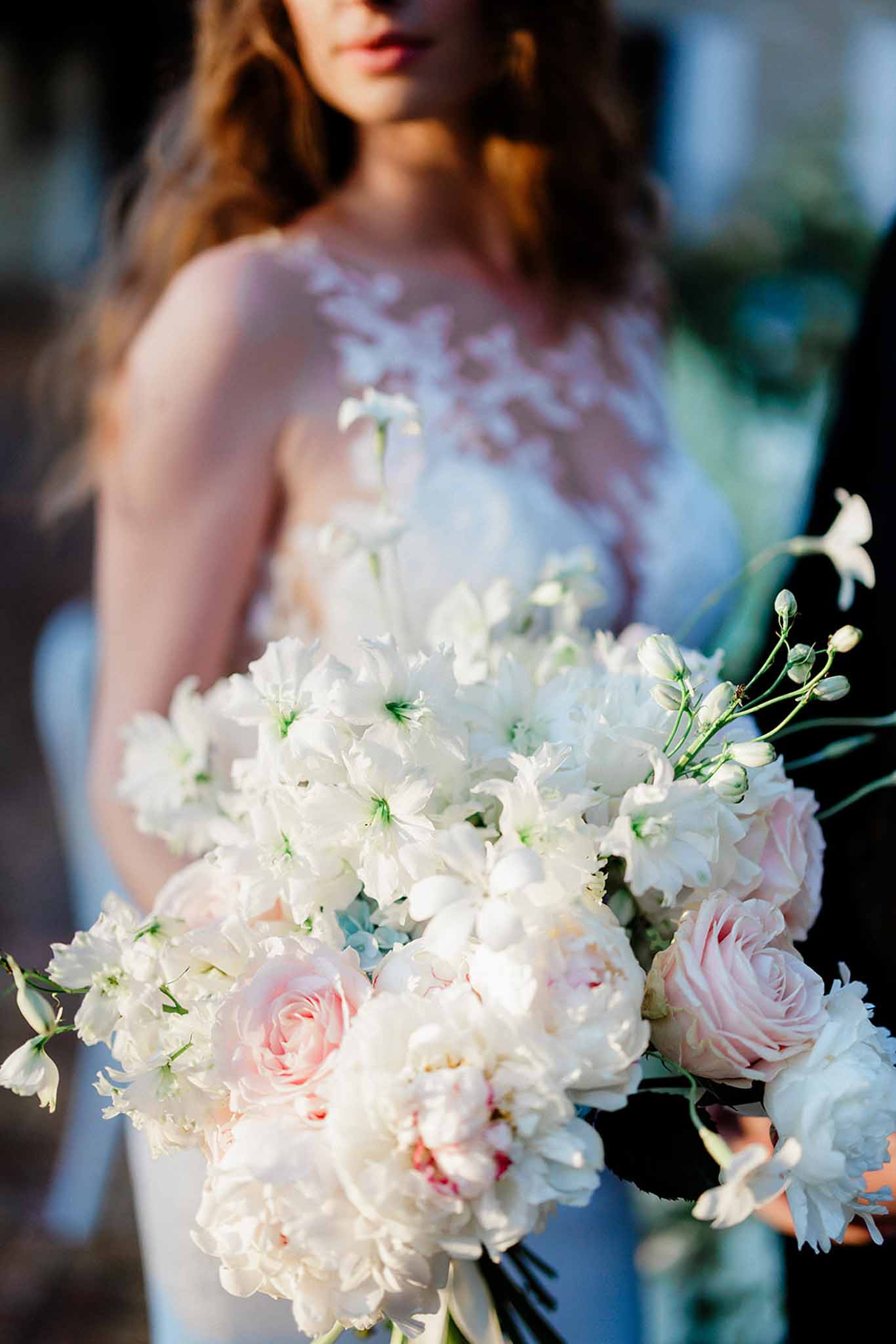 Close-up of a bridal bouquet of cream peonies, pale pink roses and baby's breath; bride in blush lace dress softly out of focus.
