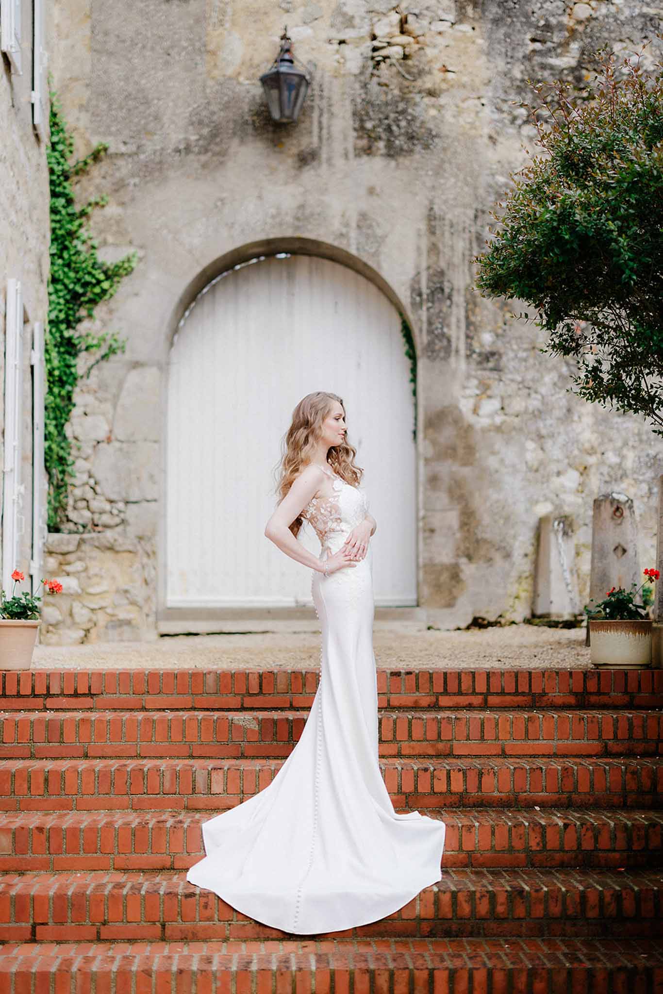 Bride in ivory column gown with long train standing in profile on red brick staircase with stone archway and climbing ivy