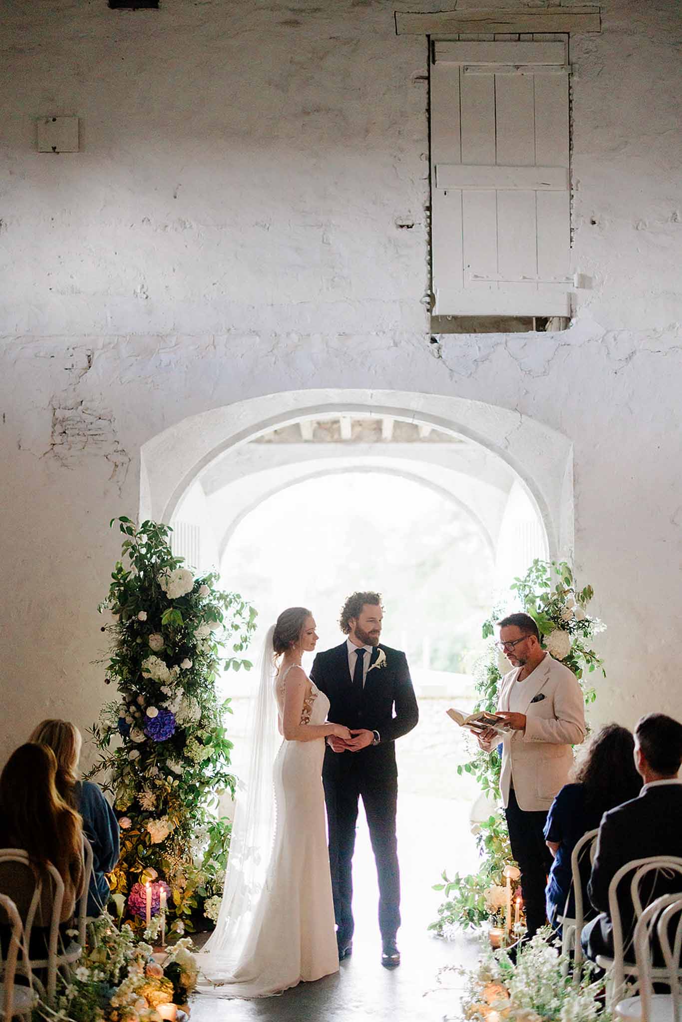 Bride and groom with officiant in whitewashed stone room with arched doorway, guests seated, floral arch behind