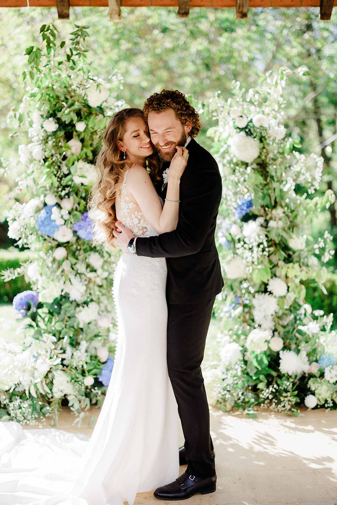 Groom lifting bride in white mermaid gown in front of white hydrangea and rose floral pergola backdrop