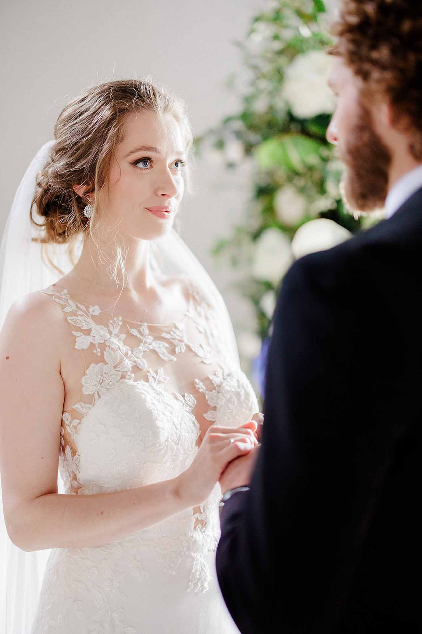 Bride in ivory lace gown with 3D floral appliqués and groom in navy suit during ceremony vows before white floral backdrop