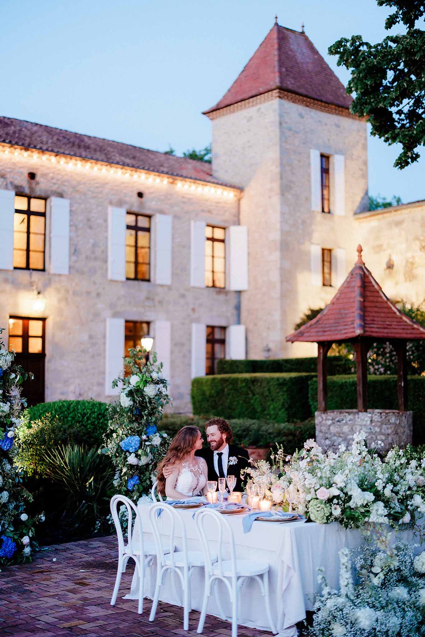 Intimate reception table for two at dusk in château courtyard with blue delphiniums and lit architectural backdrop