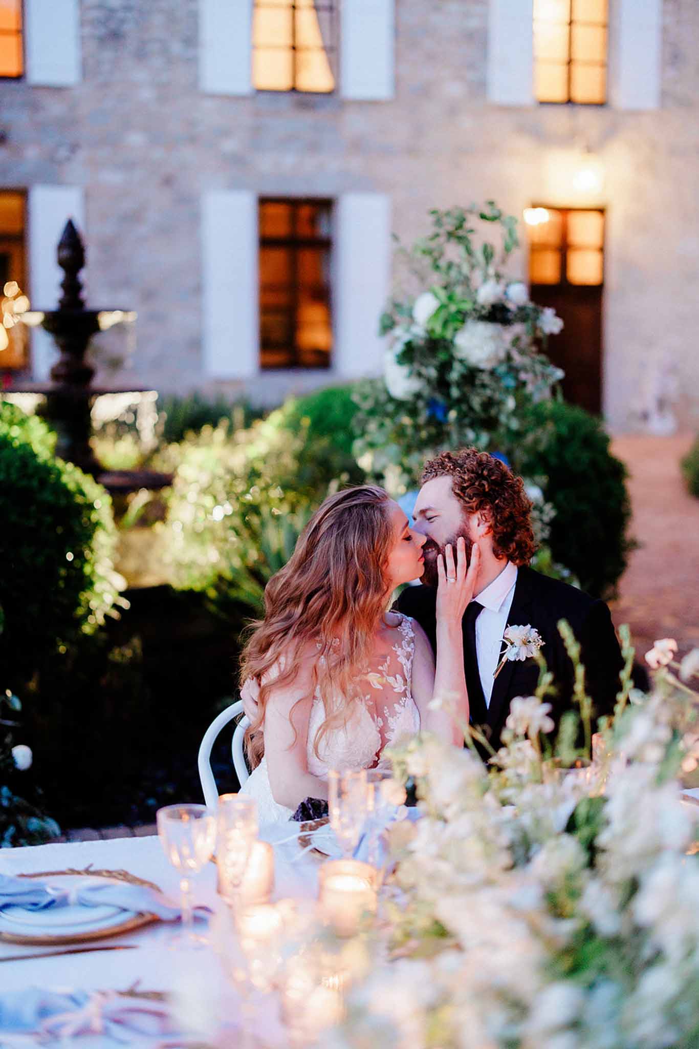 Bride and groom sharing a kiss at their candlelit reception table in an illuminated stone courtyard