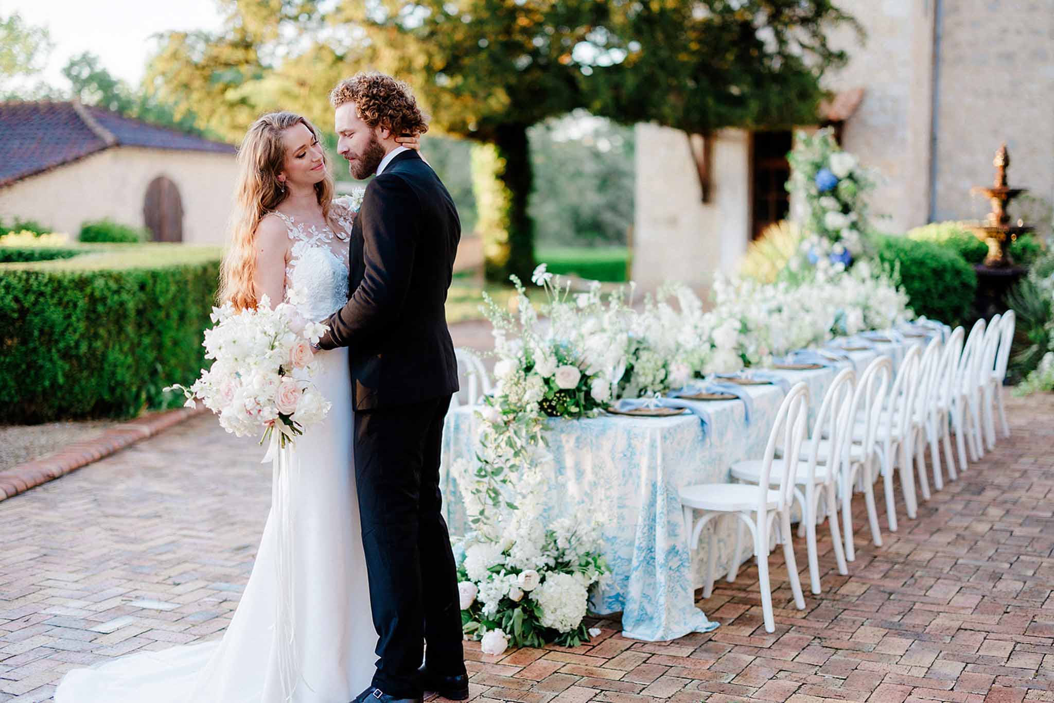 Bride in ivory lace gown with hydrangea bouquet and groom in navy suit pose before blue-cloth reception table in courtyard
