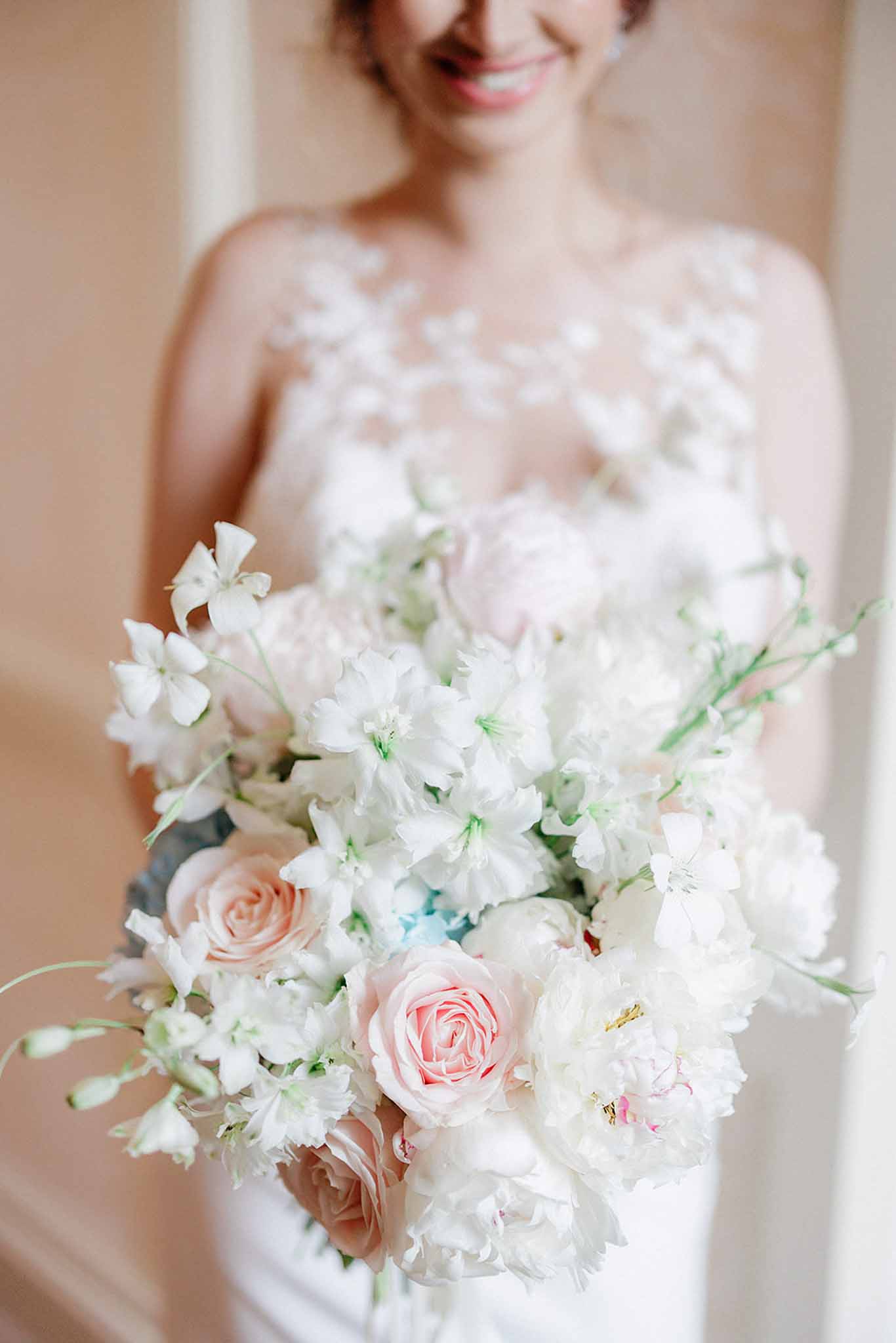 Bride in ivory lace gown with sheer sleeves holding bouquet of blush roses, white sweet peas, and green foliage against neutral wall