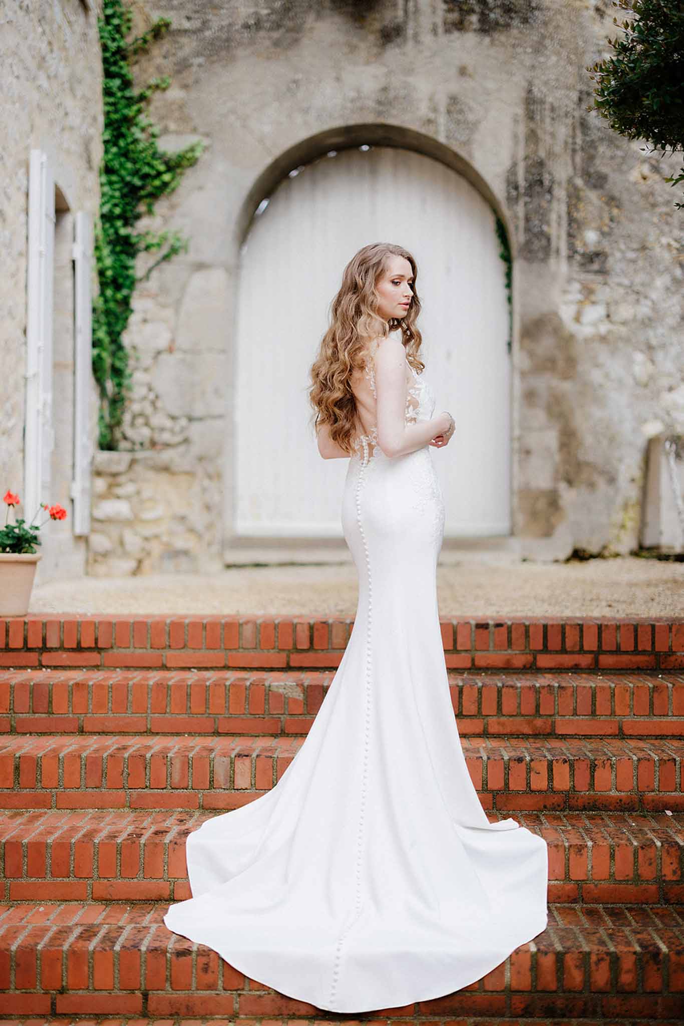 Bride in ivory mermaid gown posing in profile on red brick staircase under arched stone doorway with fairy lights