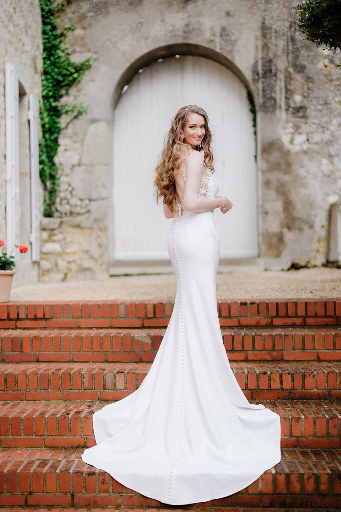 Bride in fitted white mermaid gown with lace long-sleeve bodice posing on brick staircase with ivy-covered stone walls