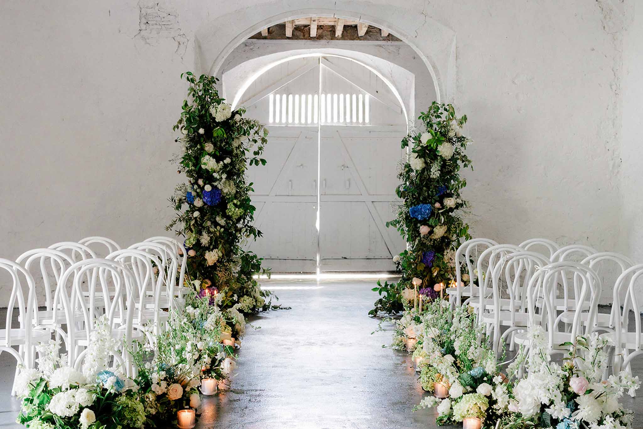 Indoor ceremony setup in vaulted industrial space with tall cascading floral installations and white bentwood chairs