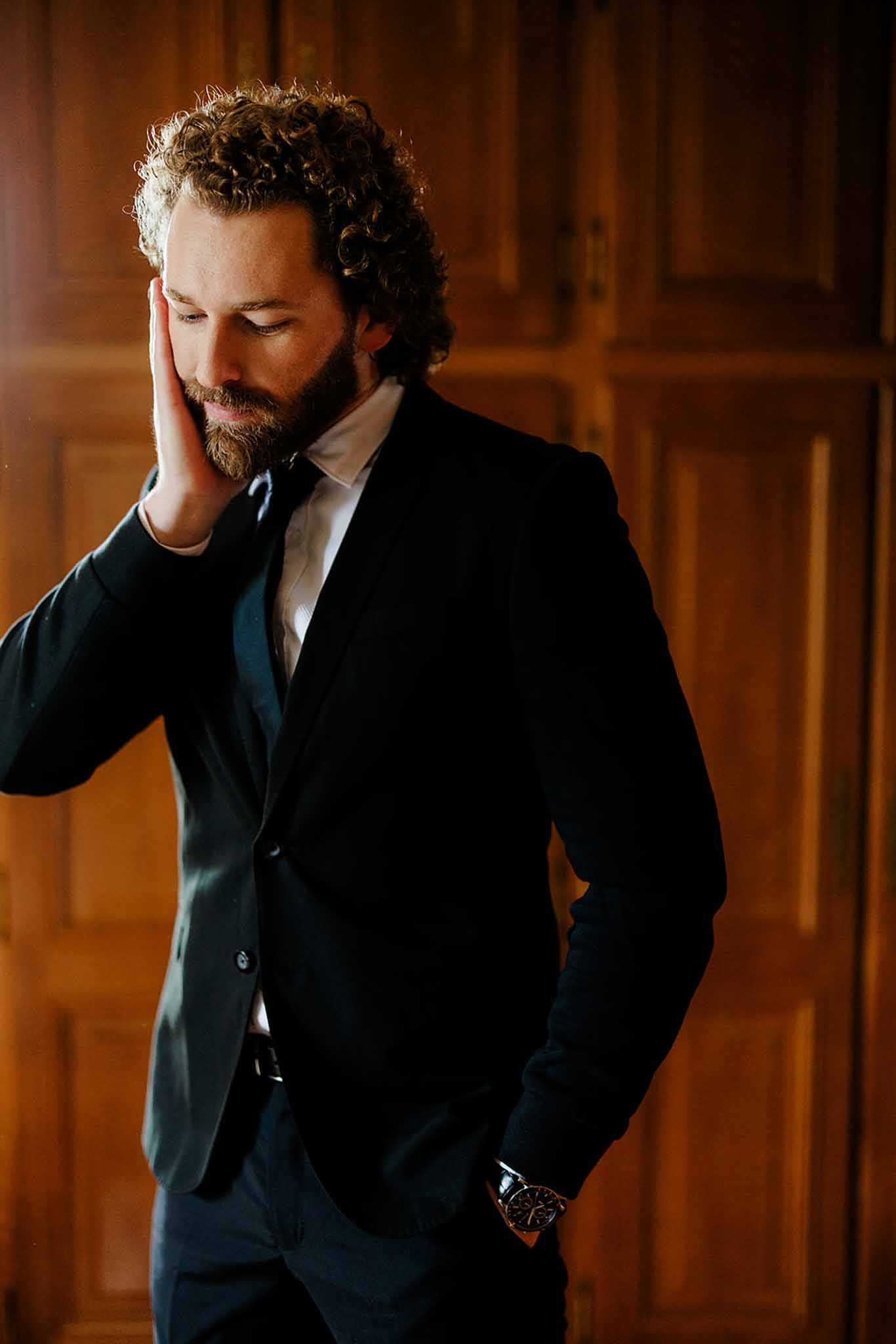 Groom in black suit with pocket square posing reflectively inside ornate wood-paneled room