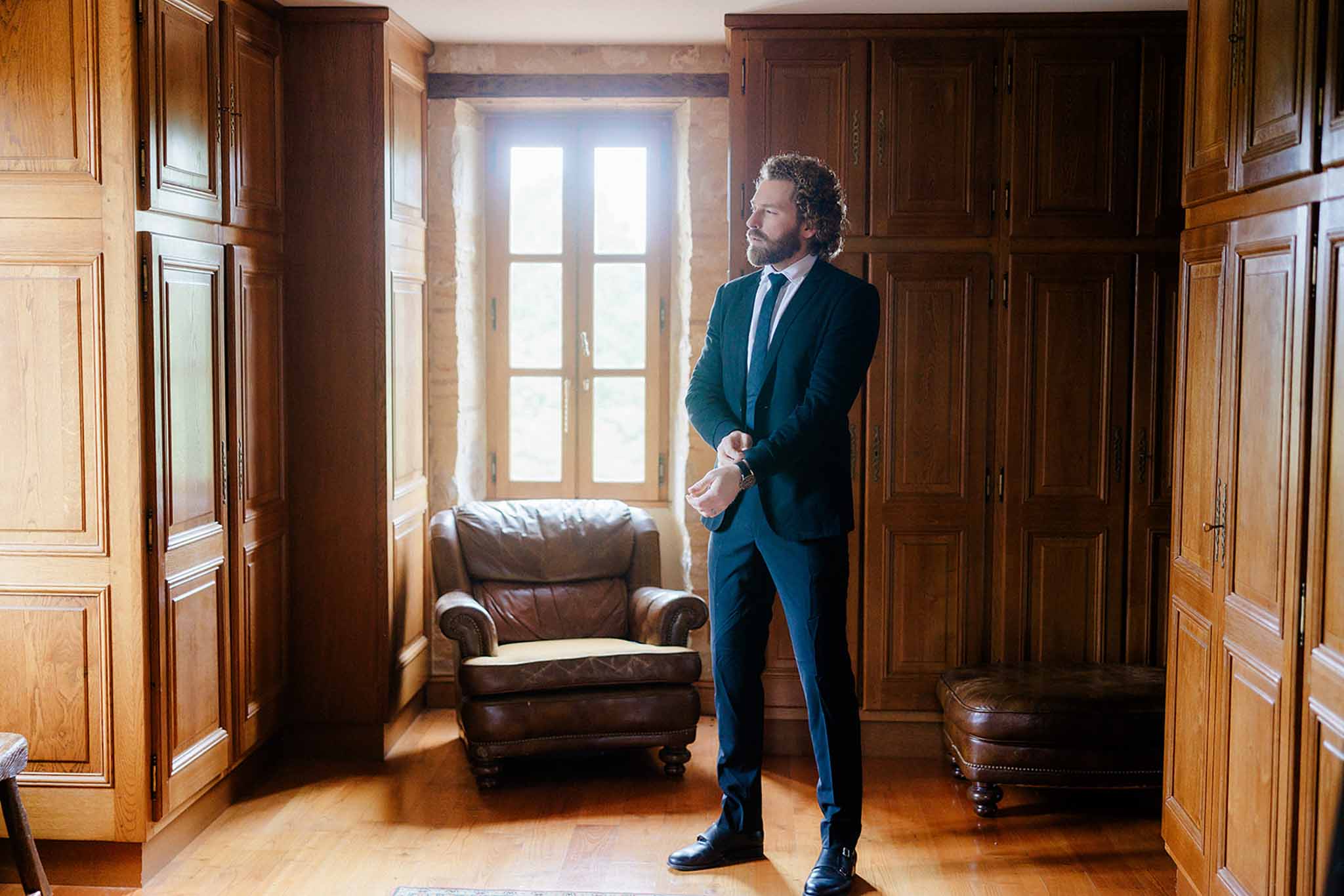 Groom in navy suit adjusting his tie in a wood-paneled hallway with leather armchair and period details
